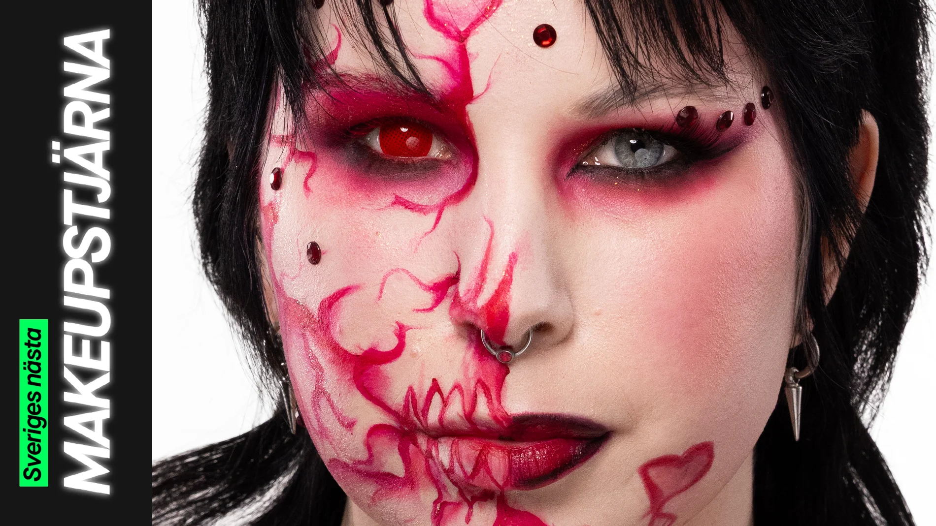 Porträtt av en person med svart hår och kreativ makeup där ansiktet är målat med röda linjer, mönster och dekorativa detaljer, fotograferad mot ljus bakgrund.