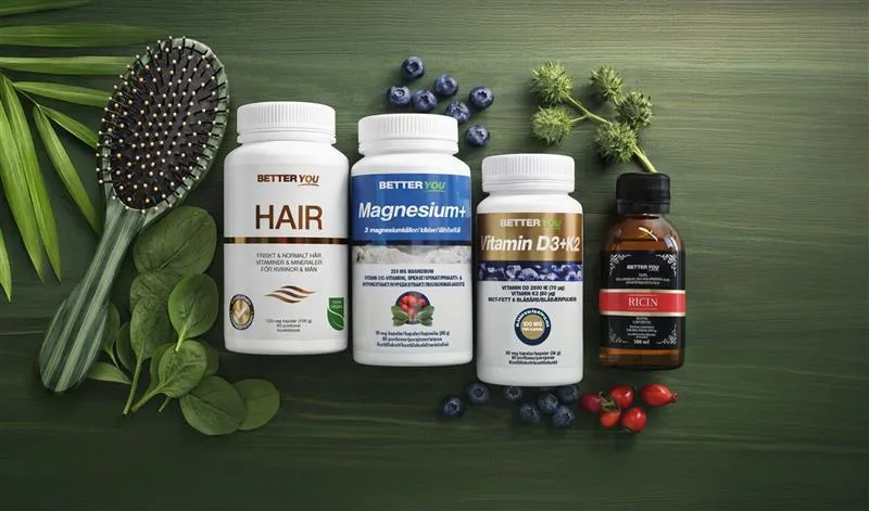 Flatlay med kosttillskott från Better You på grön bakgrund, inklusive burkar märkta “HAIR”, “Magnesium+” och “Vitamin D3+K2” samt en flaska “RICIN”. Produkterna omges av hårborste, blad, blåbär och växter som förstärker en naturlig hår- och hälsovårdskänsla.