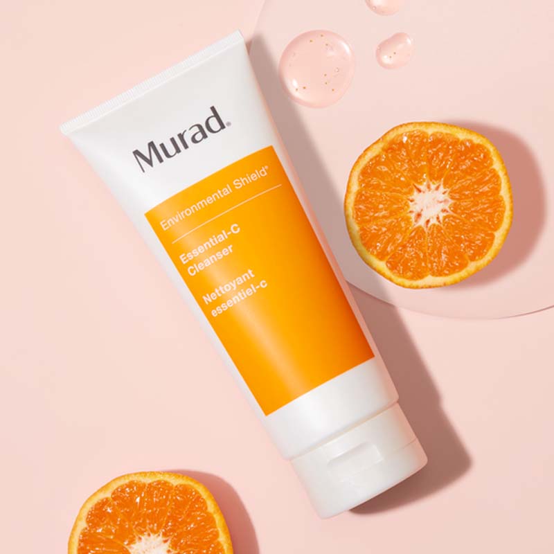 Muradin puhdistusaine vaaleanpunaisella pohjalla. Tuubi on valkoinen, ja siinä on oranssi etiketti, jossa lukee tuotteen nimi ”Environmental Shield Essential-C Cleanser”. Tuubin ympärillä on kaksi appelsiinin puolikasta ja muutama tippa tuotetta, mikä korostaa sen C-vitamiinipitoisuutta ja raikasta tunnetta.
