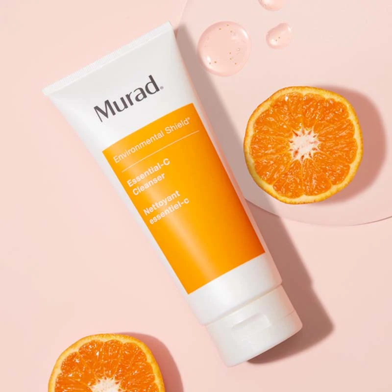 Muradin puhdistusaine vaaleanpunaisella pohjalla. Tuubi on valkoinen, ja siinä on oranssi etiketti, jossa lukee tuotteen nimi ”Environmental Shield Essential-C Cleanser”. Tuubin ympärillä on kaksi appelsiinin puolikasta ja muutama tippa tuotetta, mikä korostaa sen C-vitamiinipitoisuutta ja raikasta tunnetta.