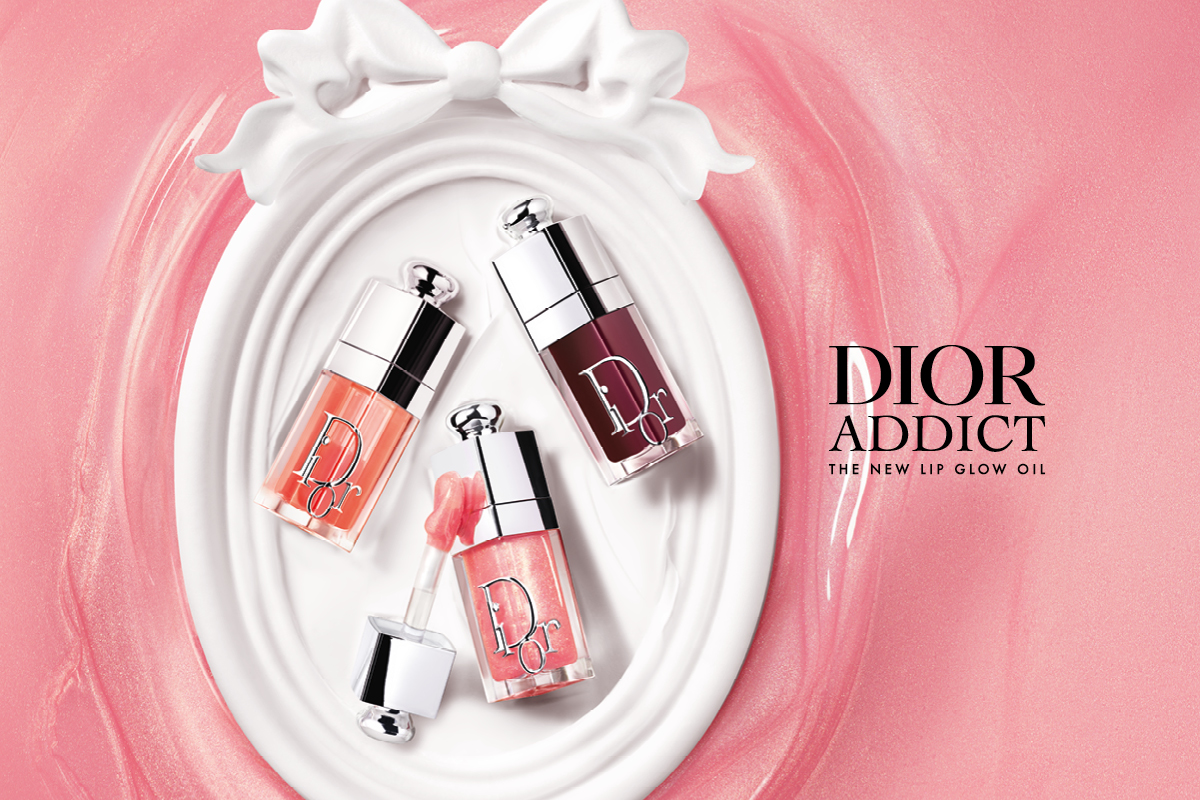 Tre Dior Addict Lip Glow Oil leppeoljer i rosa, korall og mørk nyanse med sølvlokk ligger på en hvit dekorativ overflate mot rosa bakgrunn. Tekst i bildet: "DIOR ADDICT" og "THE NEW LIP GLOW OIL".