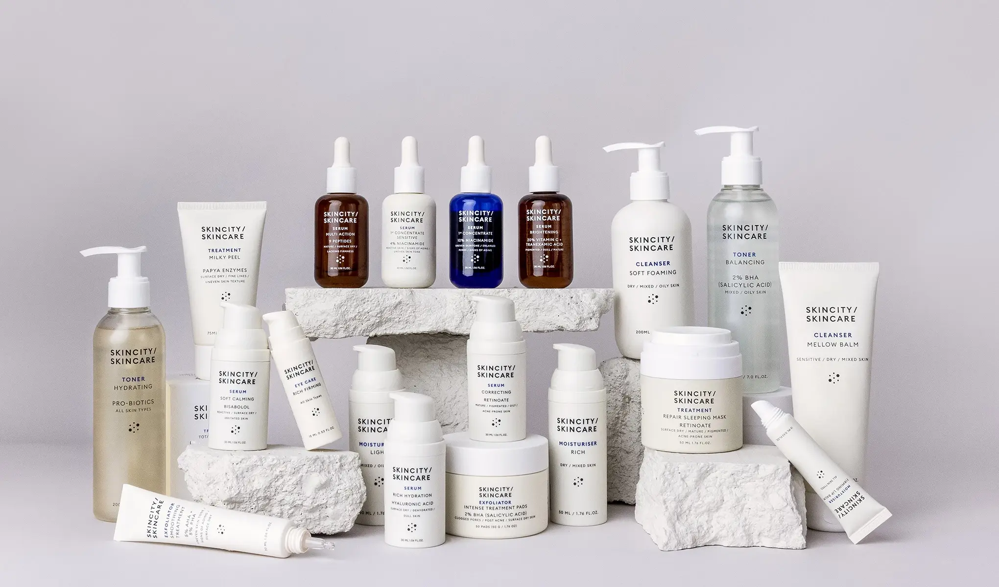 Skincity Skincare-produkter arrangert på hvite steiner mot en lysegrå bakgrunn. Flasker, krukker og tuber i nøytrale farger med minimalistisk design.