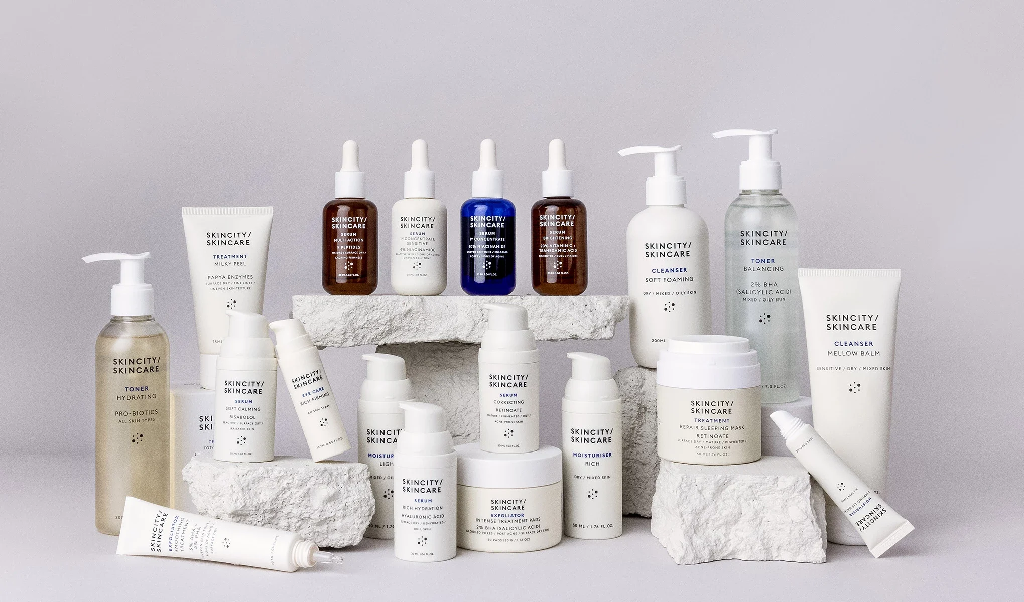Skincity Skincare-produkter arrangert på hvite steiner mot en lysegrå bakgrunn. Flasker, krukker og tuber i nøytrale farger med minimalistisk design.
