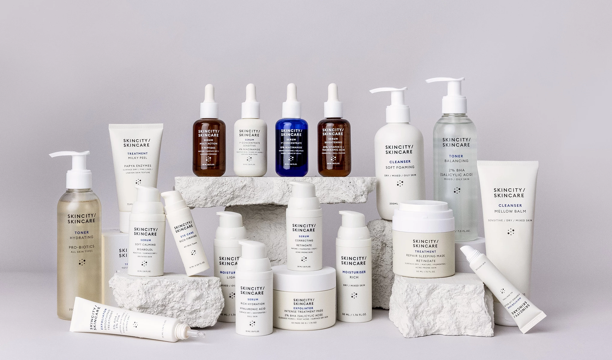 Skincity Skincare -tuotteita aseteltuna valkoisten kivien päälle vaaleanharmaata taustaa vasten. Pullot, purkit ja tuubit neutraaleissa väreissä ja minimalistisella designilla.