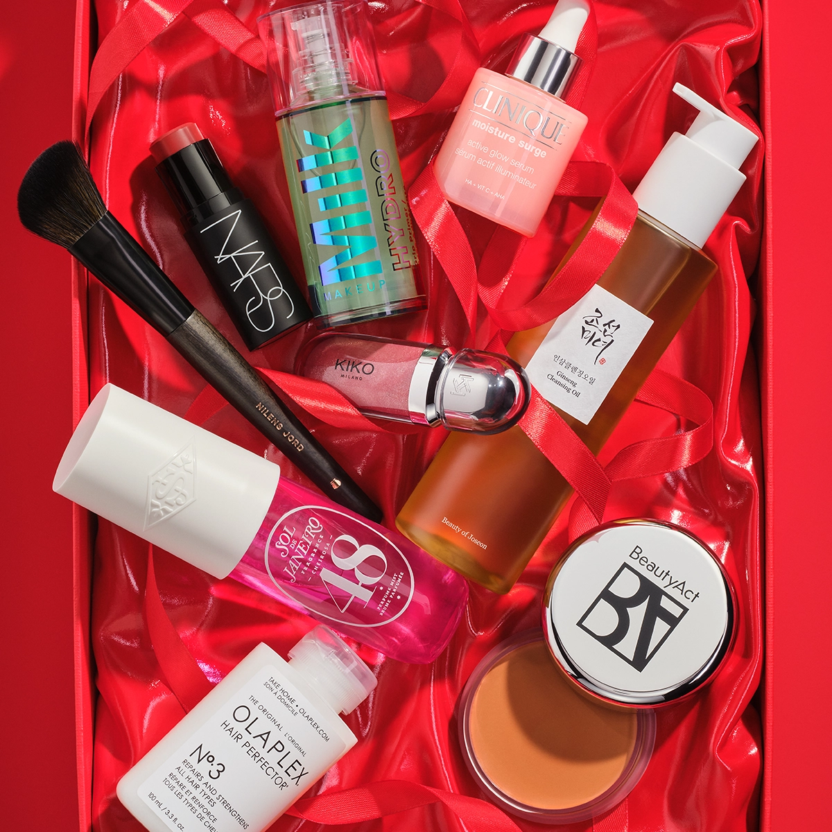 Bilde som viser flere skjønnhetsprodukter arrangert i en rød gaveeske. Produktene inkluderer en svart flaske med NARS foundation, en brun flaske med SKIN VEIL, en rosa flaske med Clinique Moisture Surge, en hvit flaske med pumpe, en boks med BeautyAct bronzer, en flaske med Olaplex Hair Perfector No.3, en sminkebørste, og flere andre skjønnhetsprodukter. Bakgrunnen er rød.
