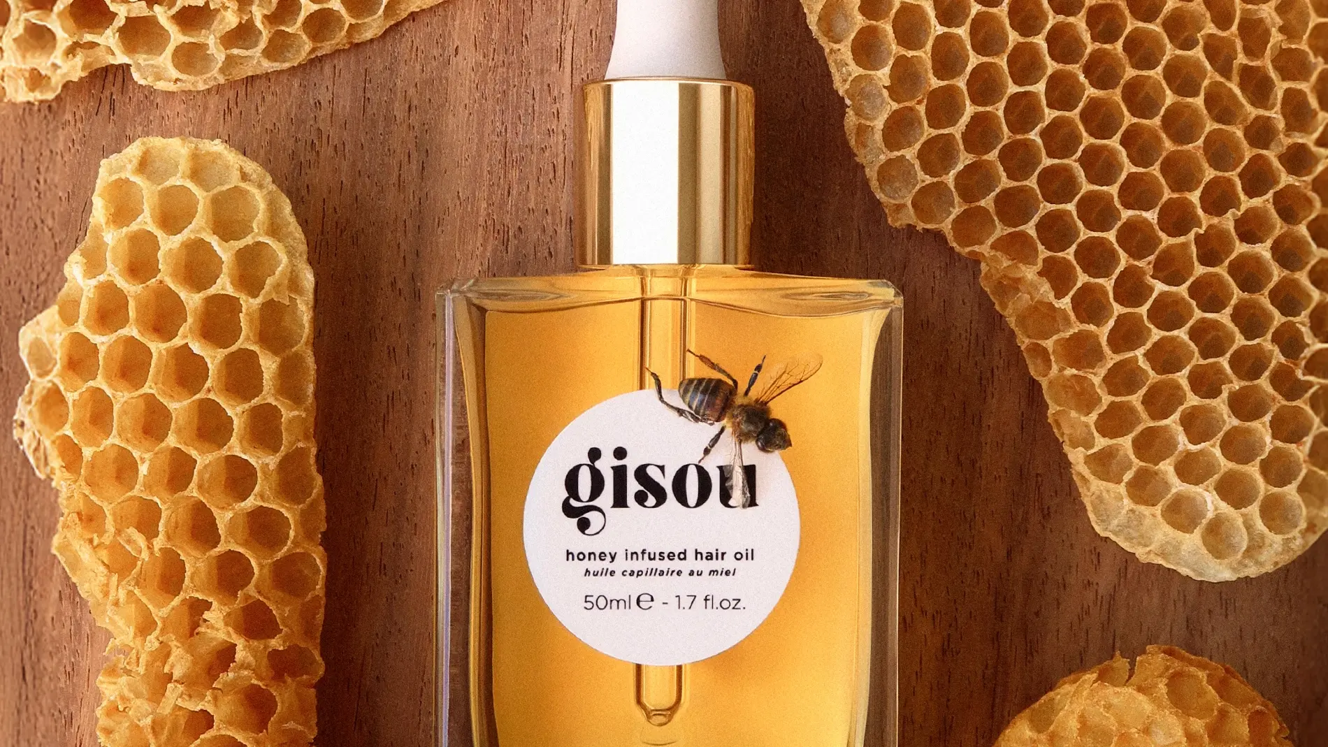 Flaske med Gisou Honey Infused Hair Oil med en bie på etiketten, omgitt av bikake. Etiketten viser teksten: 'gisou honey infused hair oil huile capillaire au miel 50ml e 1.7 fl.oz.'