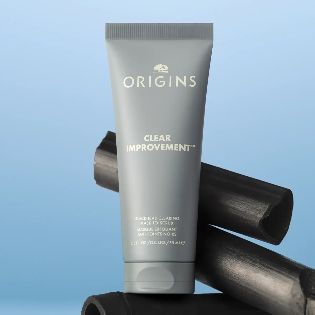Origins Clear Improvement Blackhead Clearing Mask-to-Scrub placerad framför bitar av aktivt kol mot en ljusblå bakgrund.