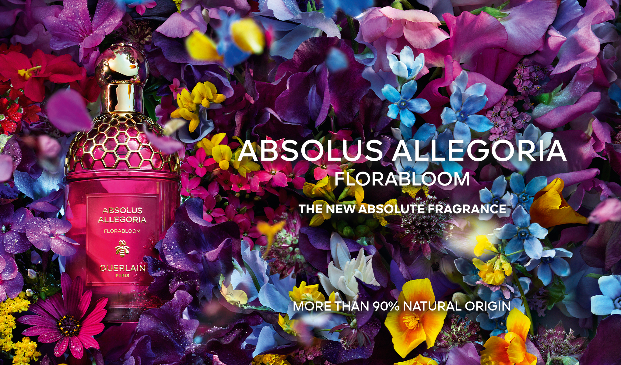 Rosa parfymeflaske med gyllen honningkakelignende kork, omgitt av fargerike blomster: lilla, blå, gule og røde blomster. Tekster: "Absolus Allegoria Florabloom", "The New Absolute Fragrance", "More than 90% natural origin".
