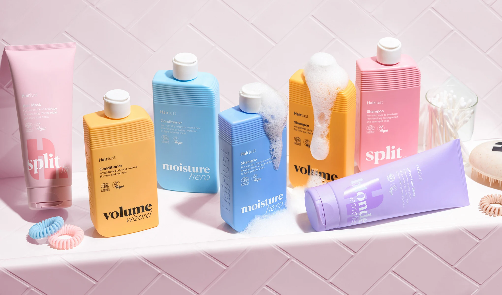 En samling hårvårdsprodukter från Hairlust är stilfullt arrangerade på en vit yta framför en rosa kaklad vägg. Produkterna är i färgglada förpackningar, såsom rosa, blå, gul och lila. Några av flaskorna har skum på sig, vilket ger en känsla av användning. Runt produkterna ligger hårsnoddar och en borste. Designen är lekfull och färgglad, med fokus på att framhäva produkternas olika funktioner och fördelar.