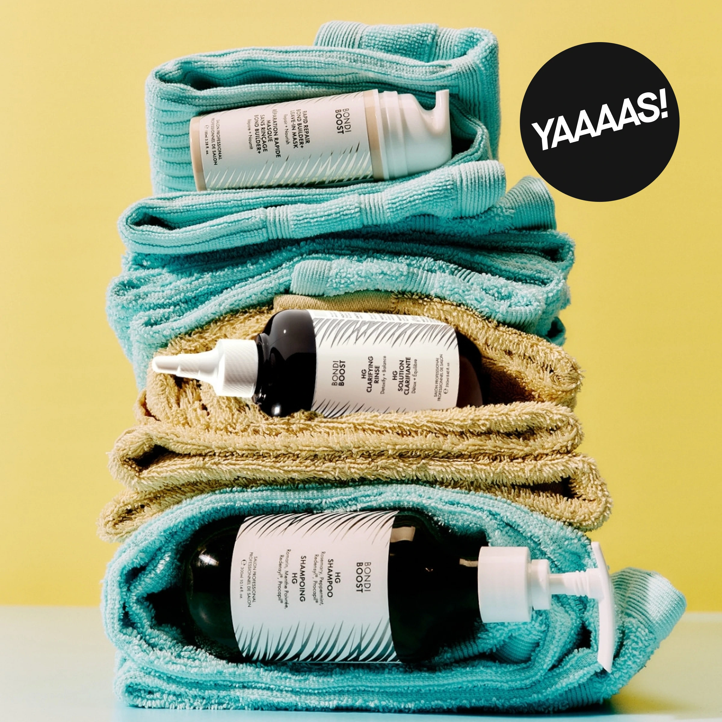 Taitellut turkoosit ja beiget pyyhkeet on pinottu Björn Axén -hiustenhoitotuotteiden kanssa, kuten “Repair Bonding Serum”, “Cleansing Conditioner” ja “Shampoo”, keltaista taustaa vasten. Mustassa ympyrässä lukee “YAAAS!”.