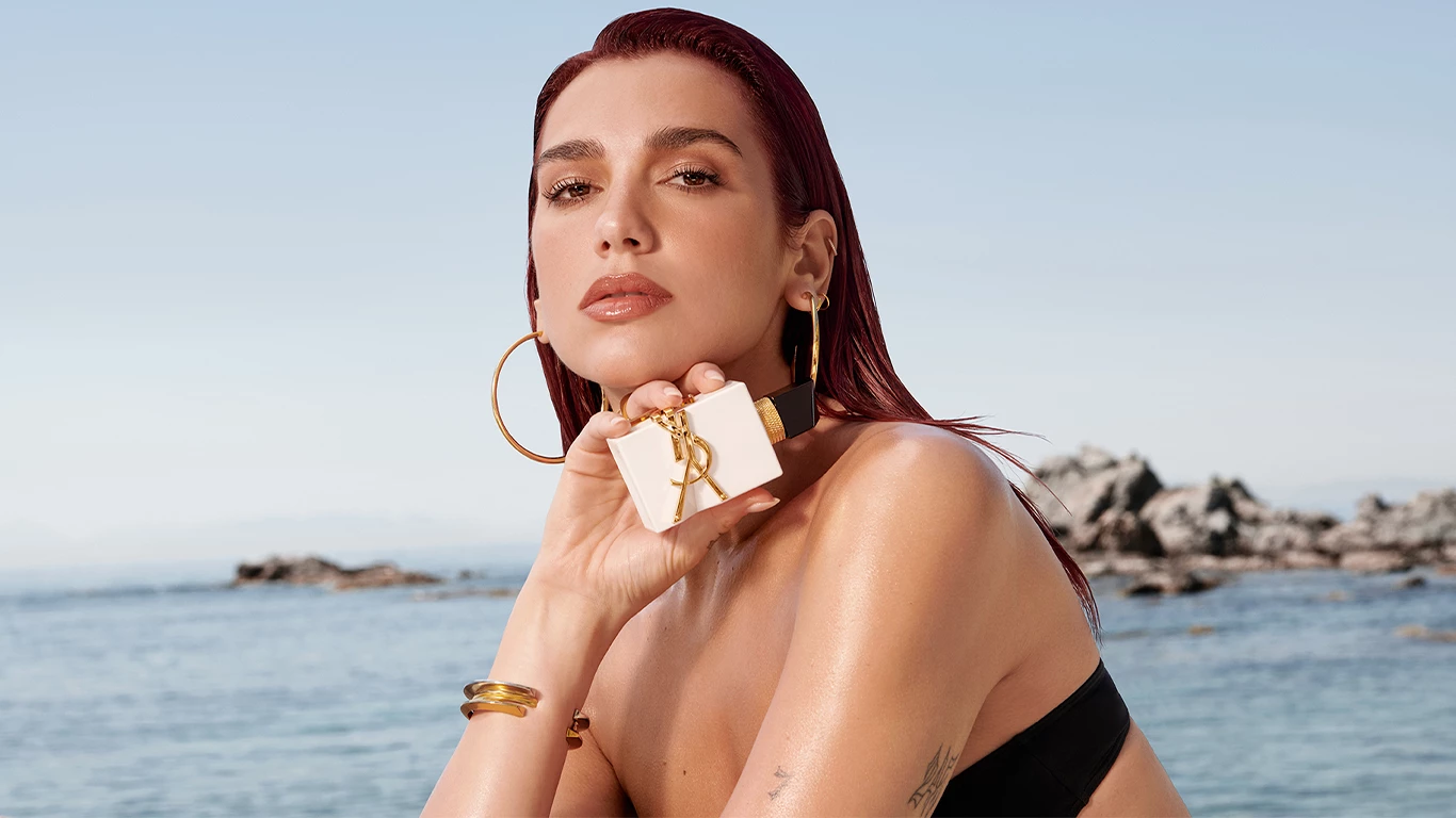 Dua Lipa håller en YSL Libre parfym i handen framfor havet.
