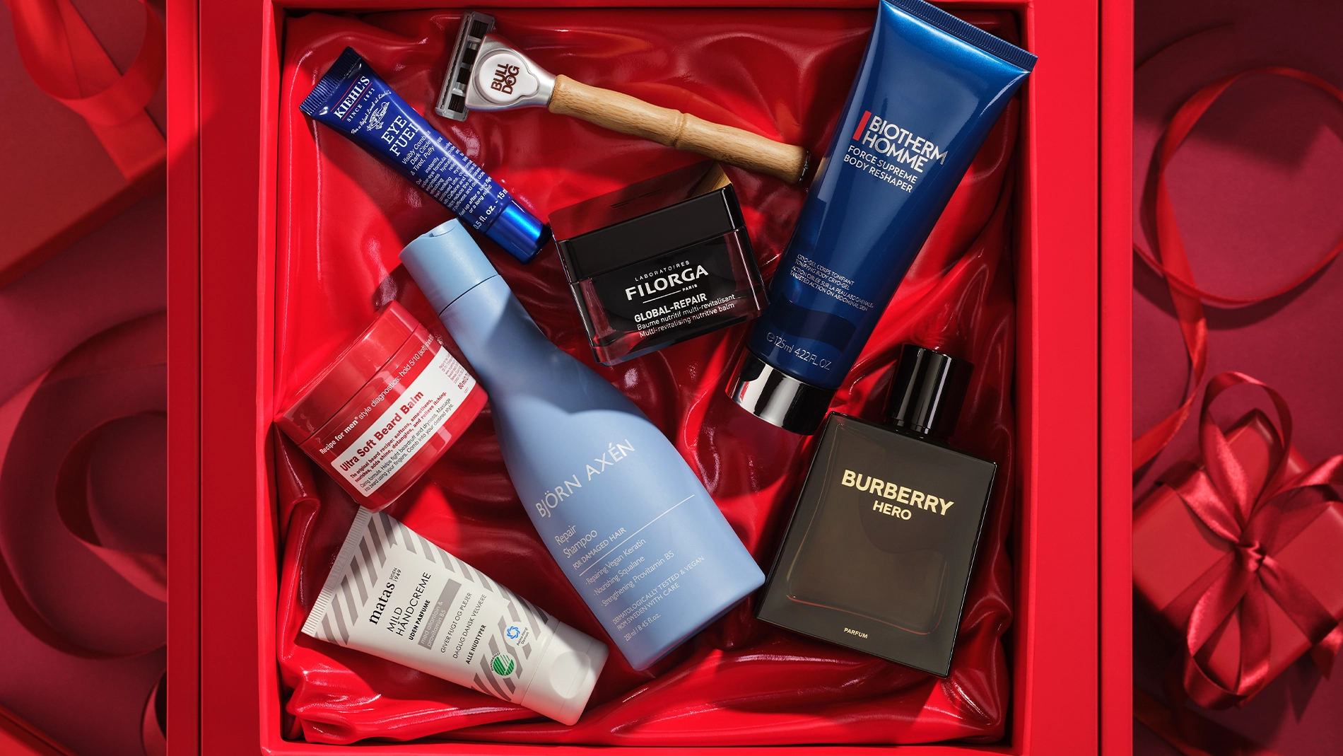 Bild som visar flera skönhetsprodukter arrangerade i en röd presentförpackning. Produkterna inkluderar en blå tub med Kiehl's Eye Fuel, en rakhyvel från Bulldog, en svart burk med Filorga Global-Repair, en blå tub med Biotherm Homme Force Supreme Body Reshaper, en svart flaska med Burberry Hero parfym, en blå flaska med Björn Axén Repair Shampoo, en vit tub med Matas Men Ansiktskräm, och en röd burk med Recipe for Men Ultra Soft Beard Balm. Bakgrunden är röd.