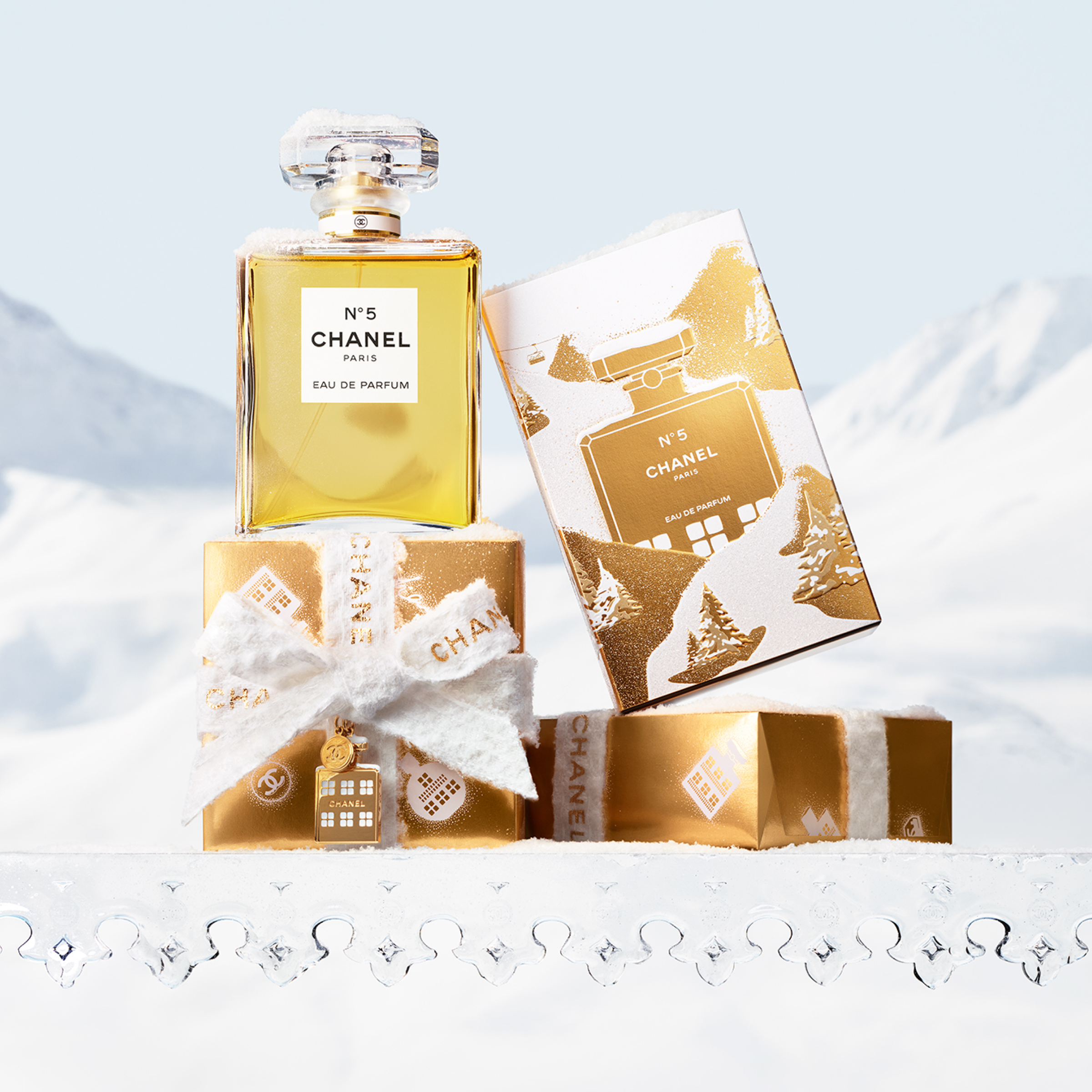 Chanel N°5 Eau de Parfum-flaske og gylne gaveesker dekket av snø, med fjellandskap i bakgrunnen. Tekst: 'N°5 CHANEL PARIS EAU DE PARFUM'.
