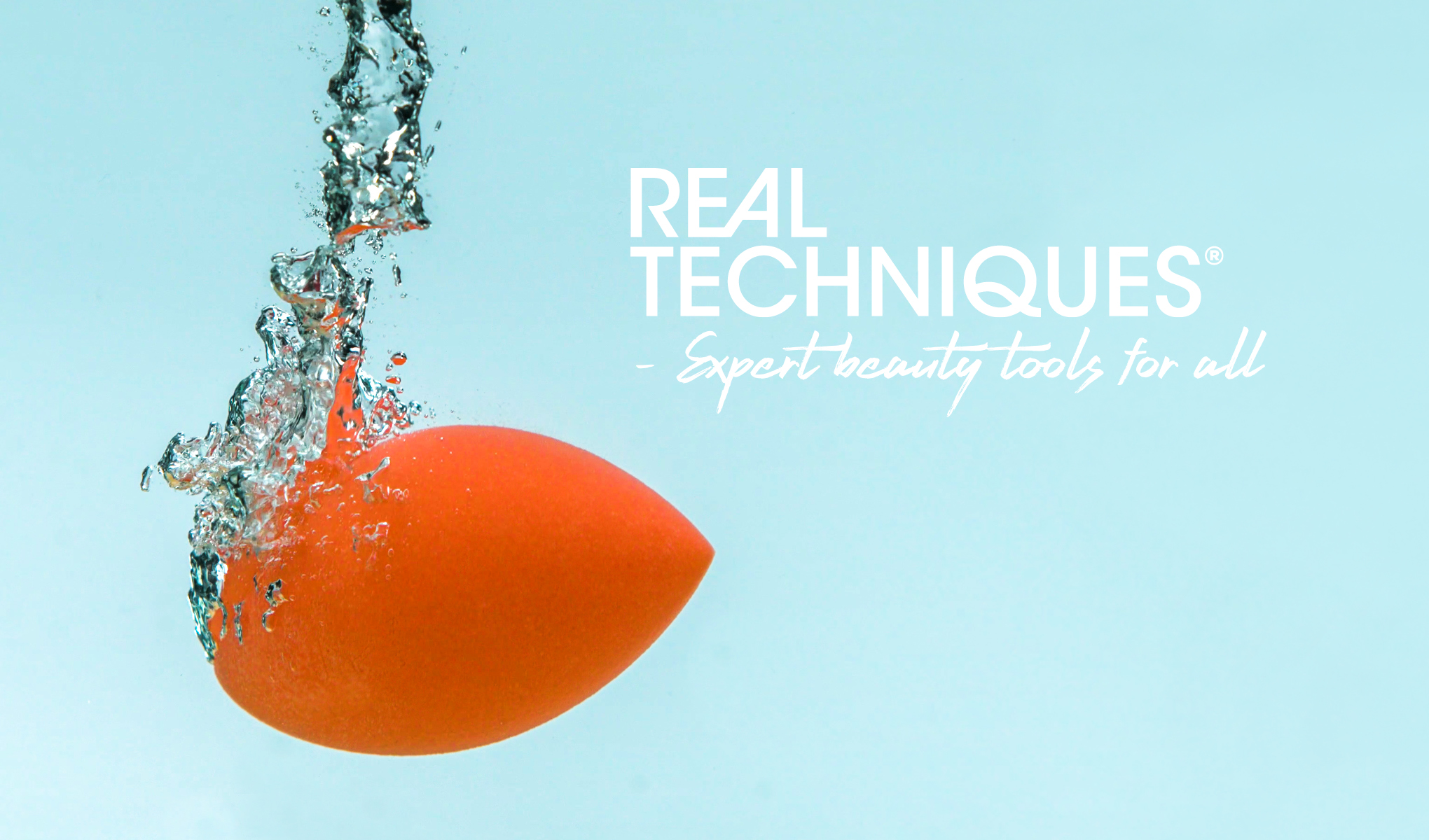 Oranssi Real Techniques -meikkisieni upotetaan veteen, jolloin syntyy roiskeita ja kuplia. Tausta on vaaleansininen, ja kuvan oikealla puolella on teksti ”REAL TECHNIQUES - Expert beauty tools for all”.