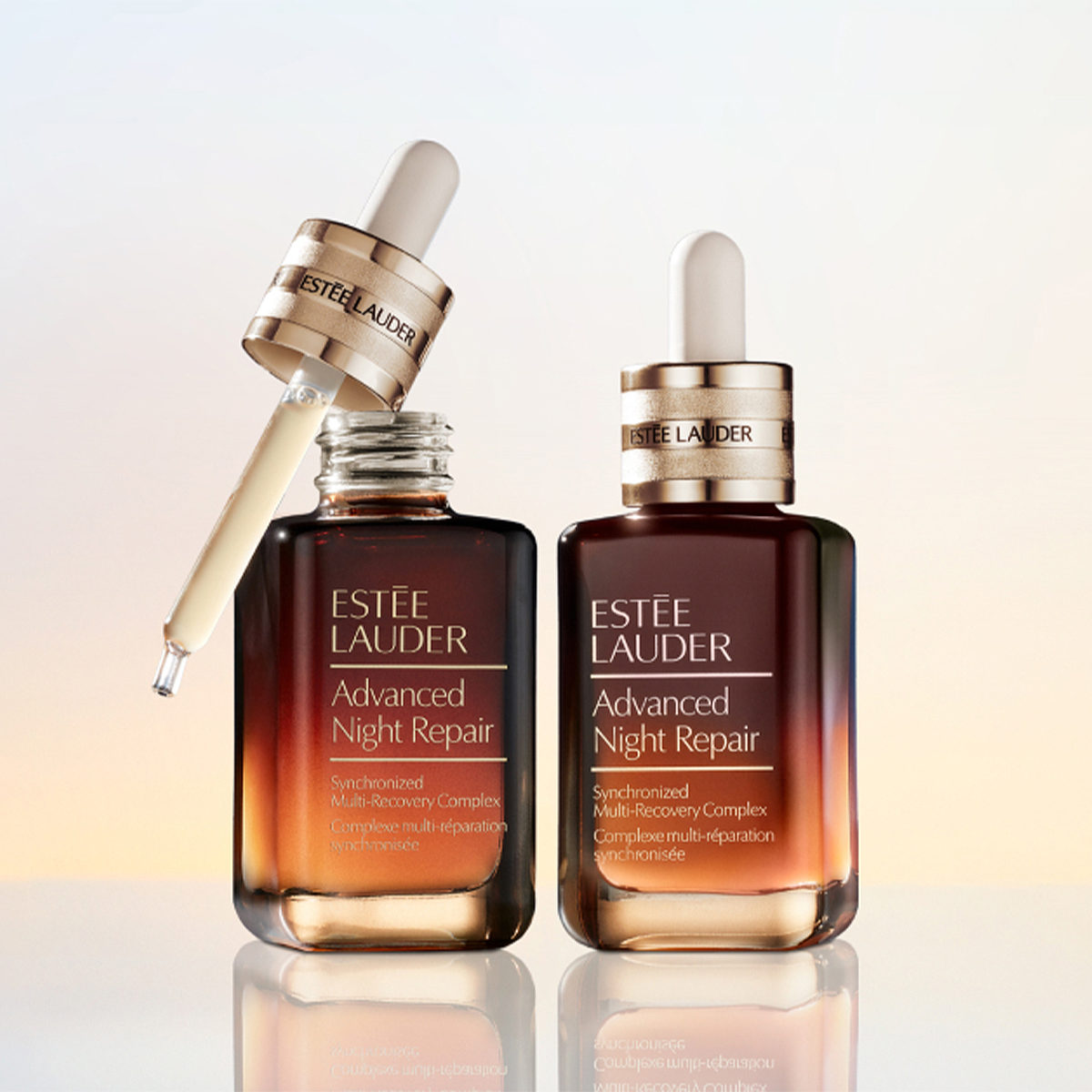 Två flaskor av Estée Lauder Advanced Night Repair serum står på en reflekterande yta mot en ljus bakgrund. Den ena flaskan är öppen med en pipett fylld med serum och visar texten ”ESTÉE LAUDER Advanced Night Repair Synchronized Multi-Recovery Complex”, medan den andra flaskan är stängd med samma produktnamn synligt.