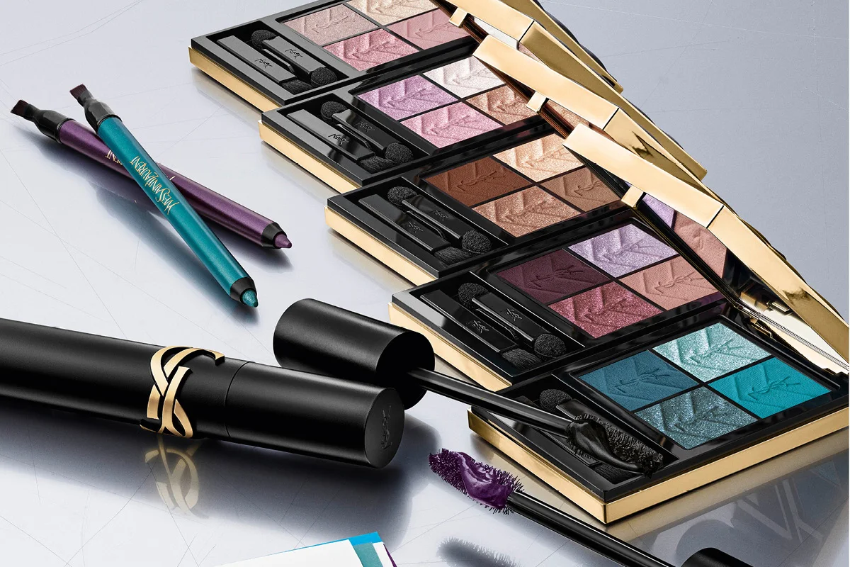 En person med lilla øyesminke holder tre sminkeprodukter fra Yves Saint Laurent Beauty foran ansiktet: en mascara i sølvfarget hylse, en lilla eyeliner og en concealer i lilla emballasje. Produktene viser YSL-logoen og teksten "Yves Saint Laurent".