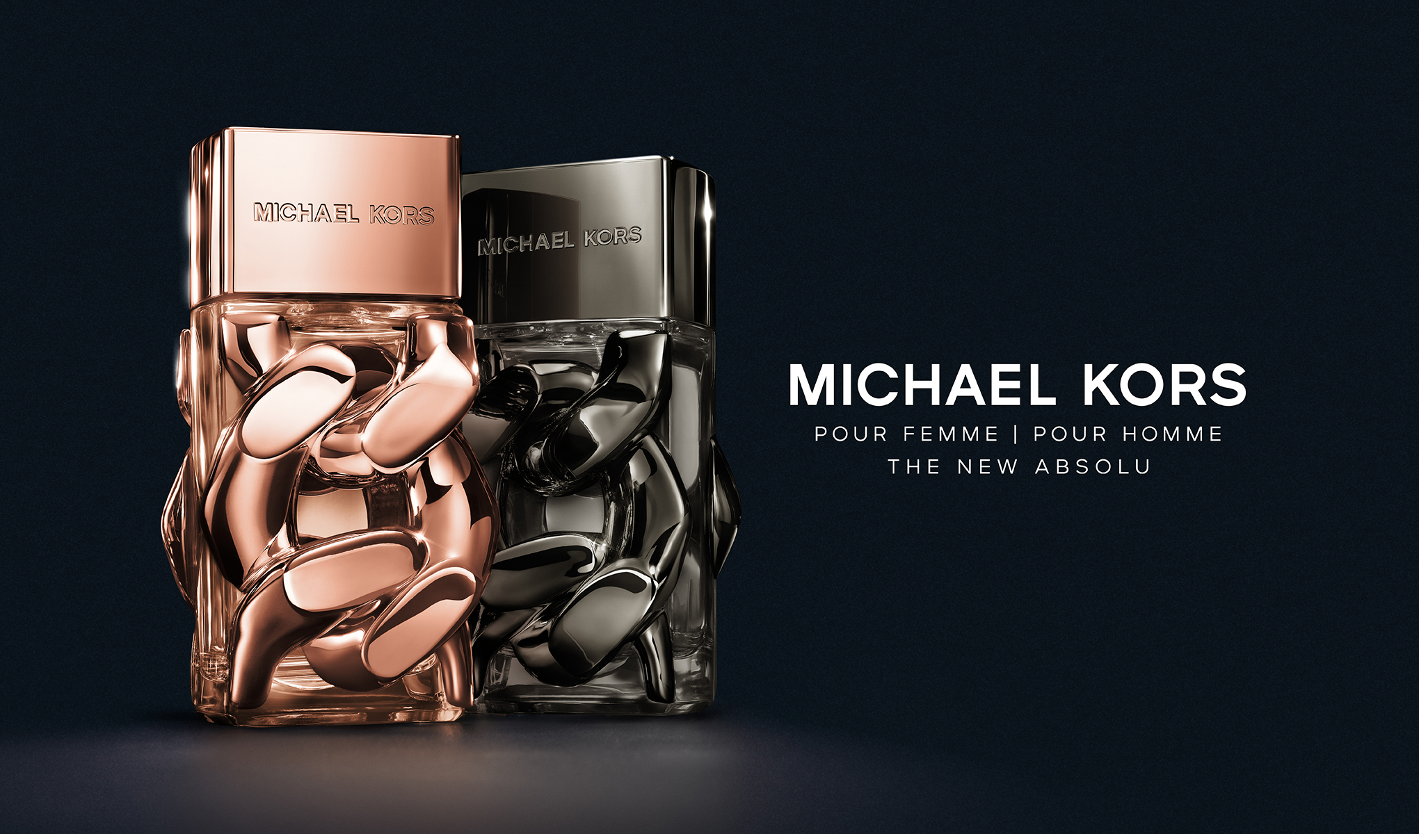 To parfymeflasker fra Michael Kors, en for kvinner og en for menn. Den kvinnelige parfymeflasken er i gull og den mannlige i sølv. Teksten på bildet lyder «Michael Kors Pour Femme | Pour Homme», og bakgrunnen er svart.