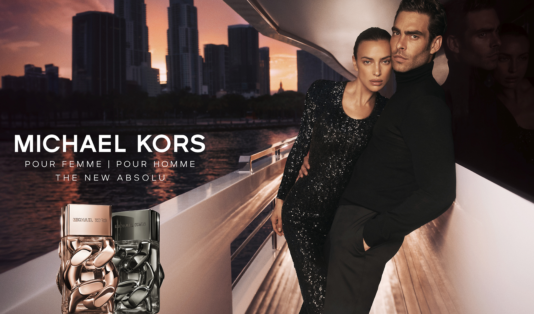 Oikealla kimaltelevaan mekkoon pukeutunut nainen ja mustaan paitaan pukeutunut mies seisovat veneessä, jonka taustalla näkyy kaupungin horisontti auringonlaskun aikaan. Vasemmalla etualalla kaksi hajuvesipulloa, toinen naisille ja toinen miehille. Kuvassa on teksti ”Michael Kors Pour Femme | Pour Homme”.