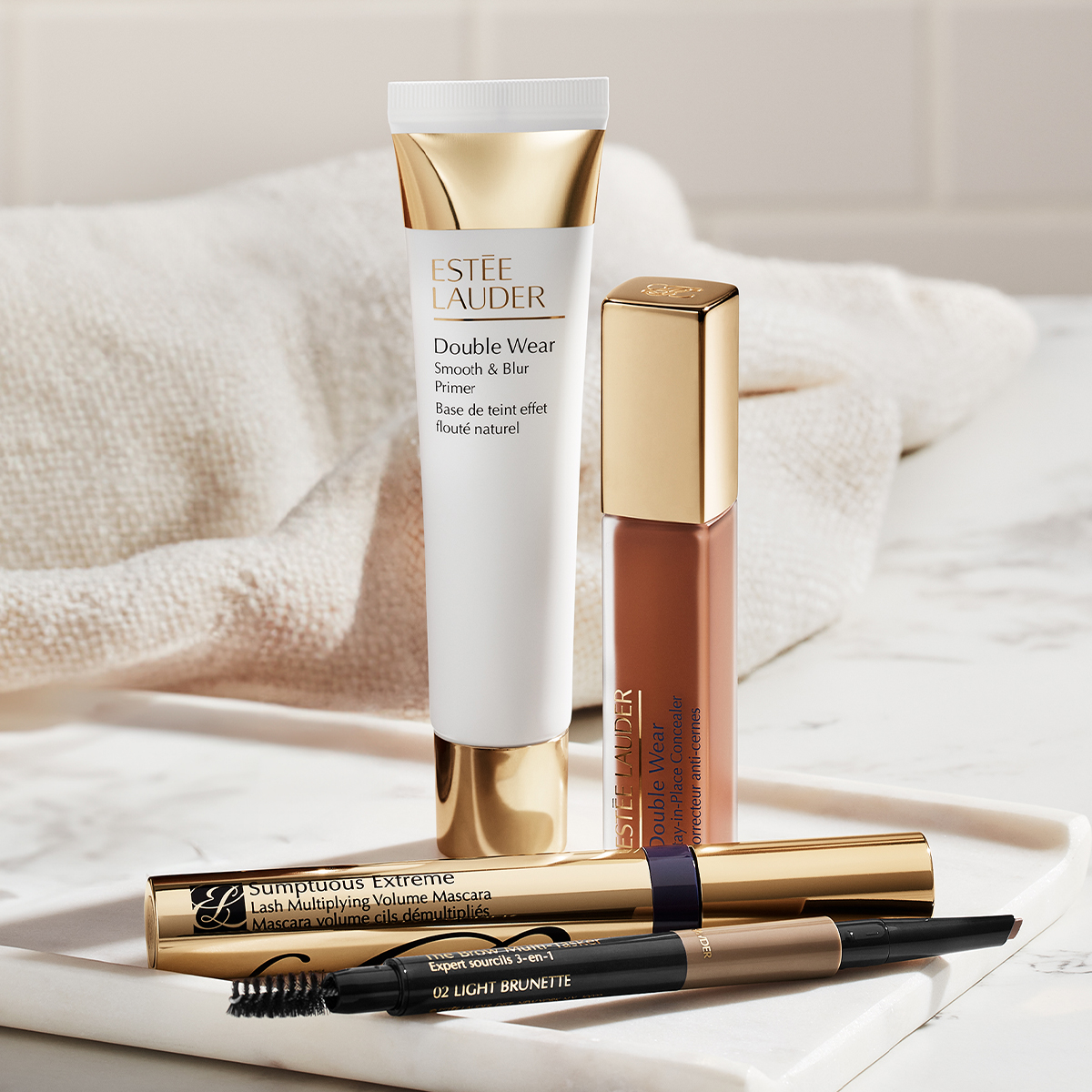Utvalda sminkprodukter från Estée Lauder på marmoryta: Double Wear Smooth & Blur primer, Double Wear concealer, mascara och ögonbrynsprodukt mot en mjuk tygbakgrund.