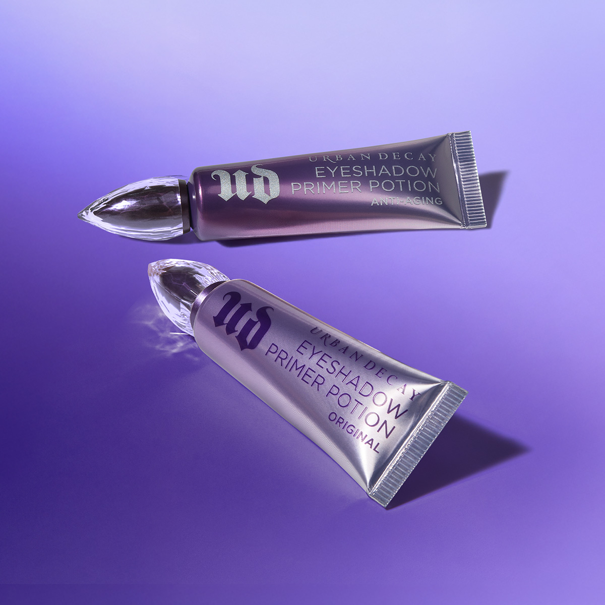 Två lila tuber med texterna "URBAN DECAY EYESHADOW PRIMER POTION ORIGINAL" och "URBAN DECAY EYESHADOW PRIMER POTION ANTI-AGING" ligger på en lila bakgrund.