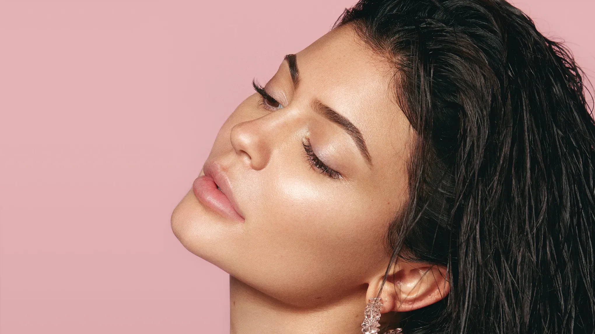 Kylie Jenner profiilissa, katsoo alas ja kallistaa päätään taaksepäin vaaleanpunaista taustaa vasten.