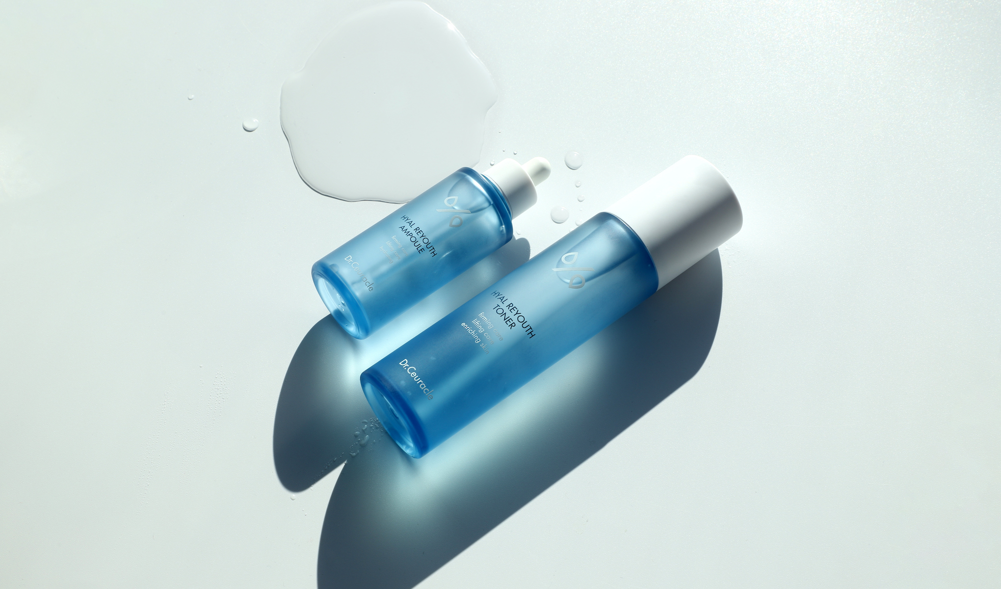 To blå flasker med Dr. Ceuracle Hyal Reyouth Toner og Hyal Reyouth Ampoule, plassert på en hvit flate med noen dråper vann.