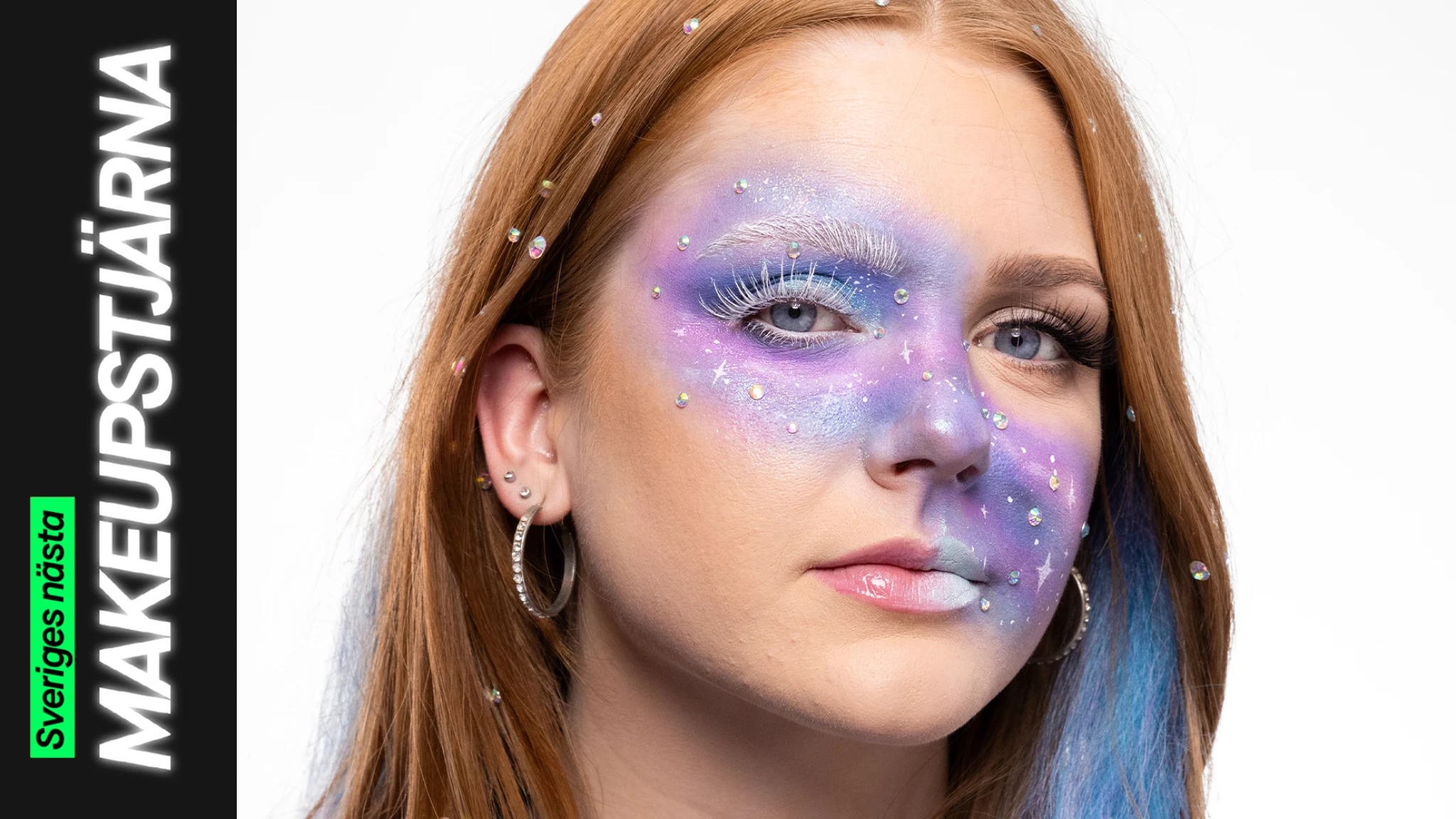 En person med galaxinspirerad makeup i lila, blå och silvertoner med vita fransar, små glittrande stenar och stjärnliknande detaljer över ansiktet och i håret.