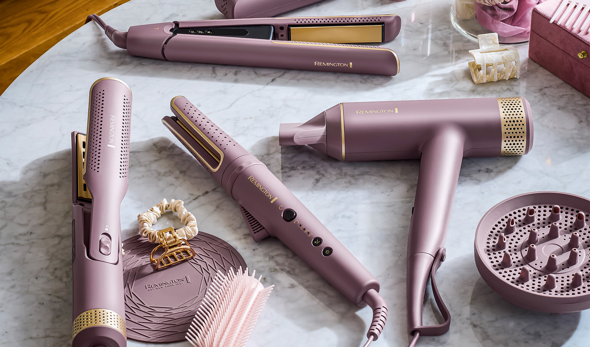 En samling hårstylingverktøy, inkludert en hårføner, en rettetang og to elektriske barbermaskiner, er elegant arrangert på en mørk overflate med en minimalistisk bakgrunn. Verktøyene har en moderne design med svarte og bronsefargede detaljer.