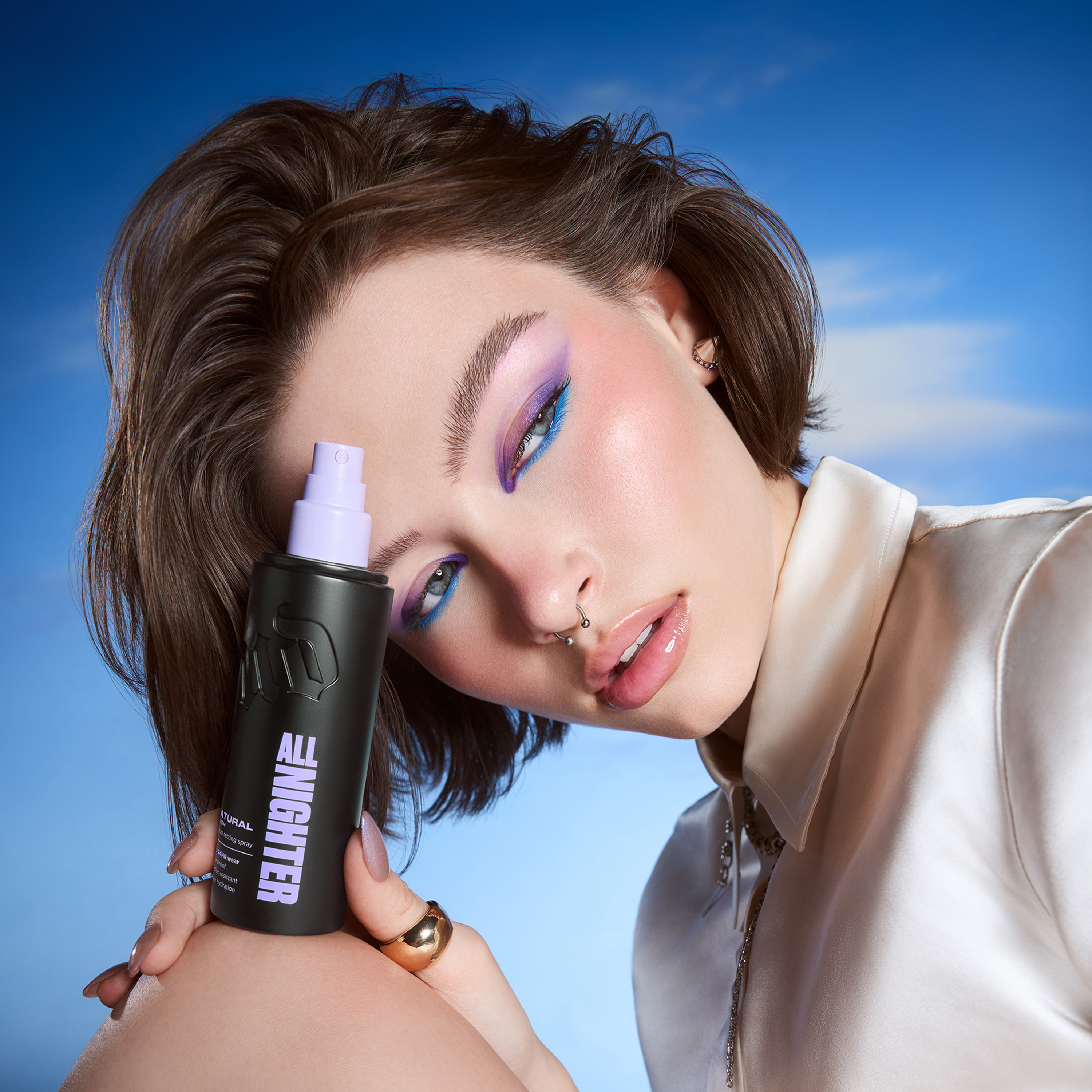 Person med färgstark ögonmakeup i lila och blått håller Urban Decay All Nighter setting spray i svart flaska med lila spraypump mot blå bakgrund. På flaskan syns texten “ALL NIGHTER”.
