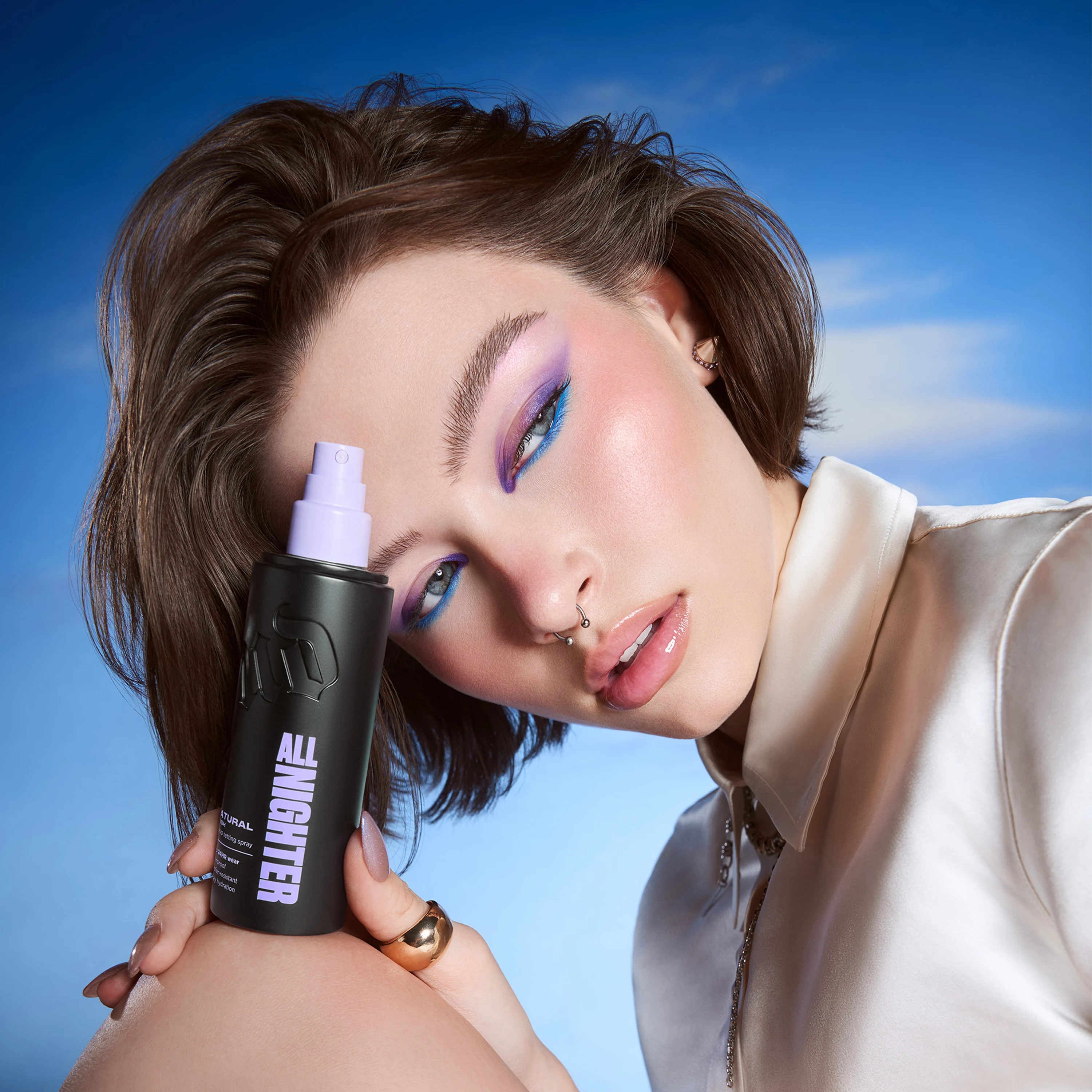 Person med färgstark ögonmakeup i lila och blått håller Urban Decay All Nighter setting spray i svart flaska med lila spraypump mot blå bakgrund. På flaskan syns texten “ALL NIGHTER”.