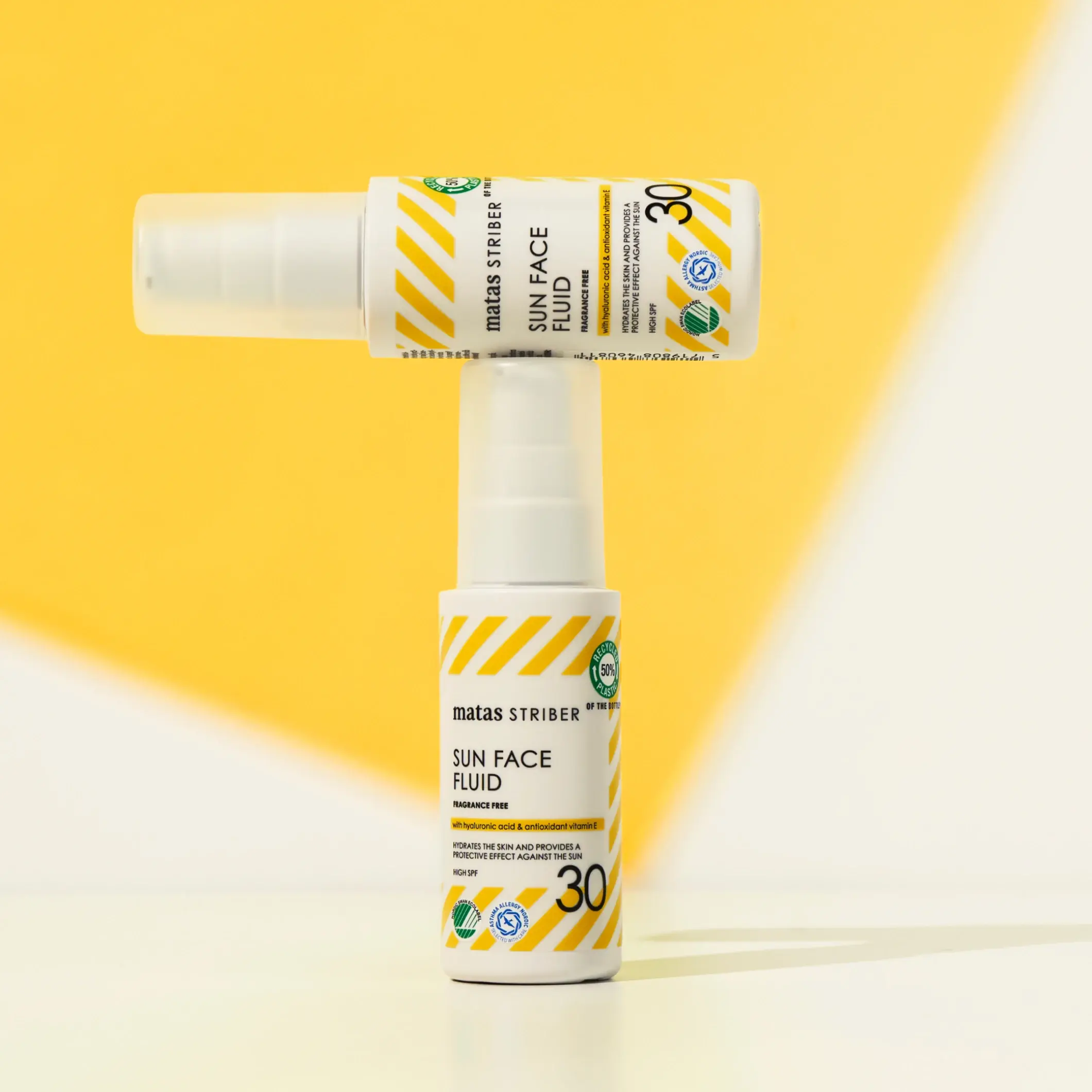 To Matas Striber Sun Face Fluid SPF 30 solbeskyttelsesflasker, hvor den ene ligger horisontalt oppå den andre mot en gul bakgrunn.