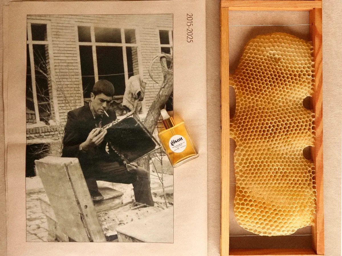 Mustavalkokuva tupakoivasta miehestä, Gisou Honey Infused Hair Oil -pullo ja hunajakenno puukehyksessä. Teksti '2015-2025'.