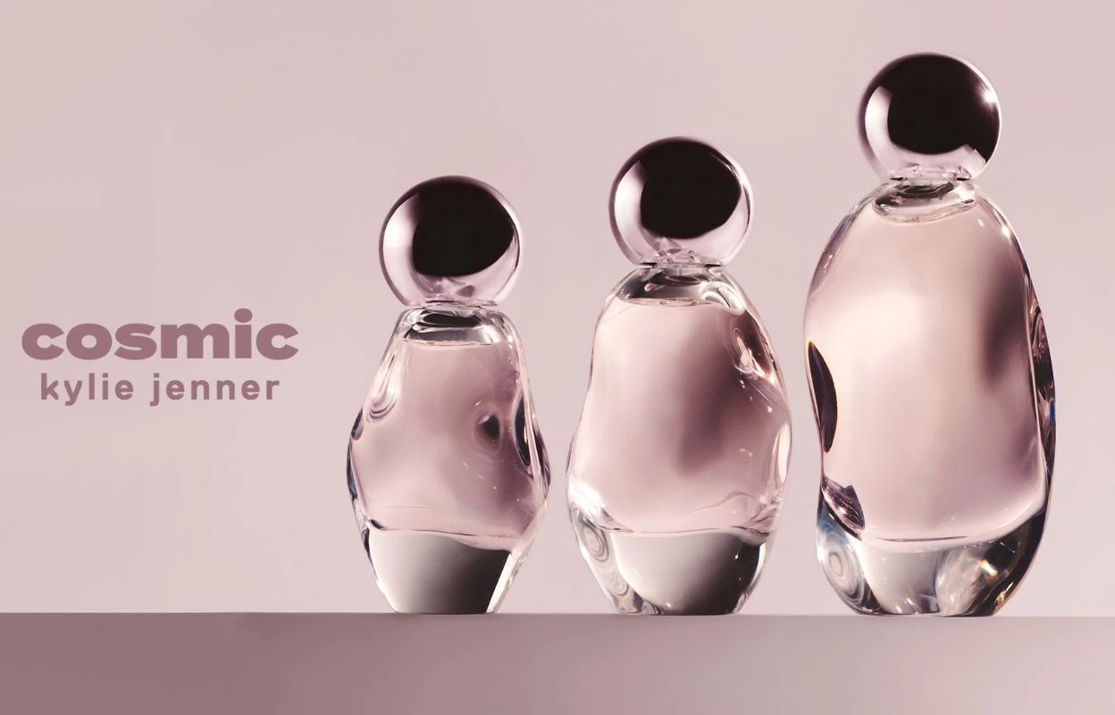 Kolme Kylie Cosmeticsin hajuvettä vaaleanpunaisella taustalla, yhdessä tummanvaaleanpunaisen tekstin "cosmic kylie jenner" kanssa.