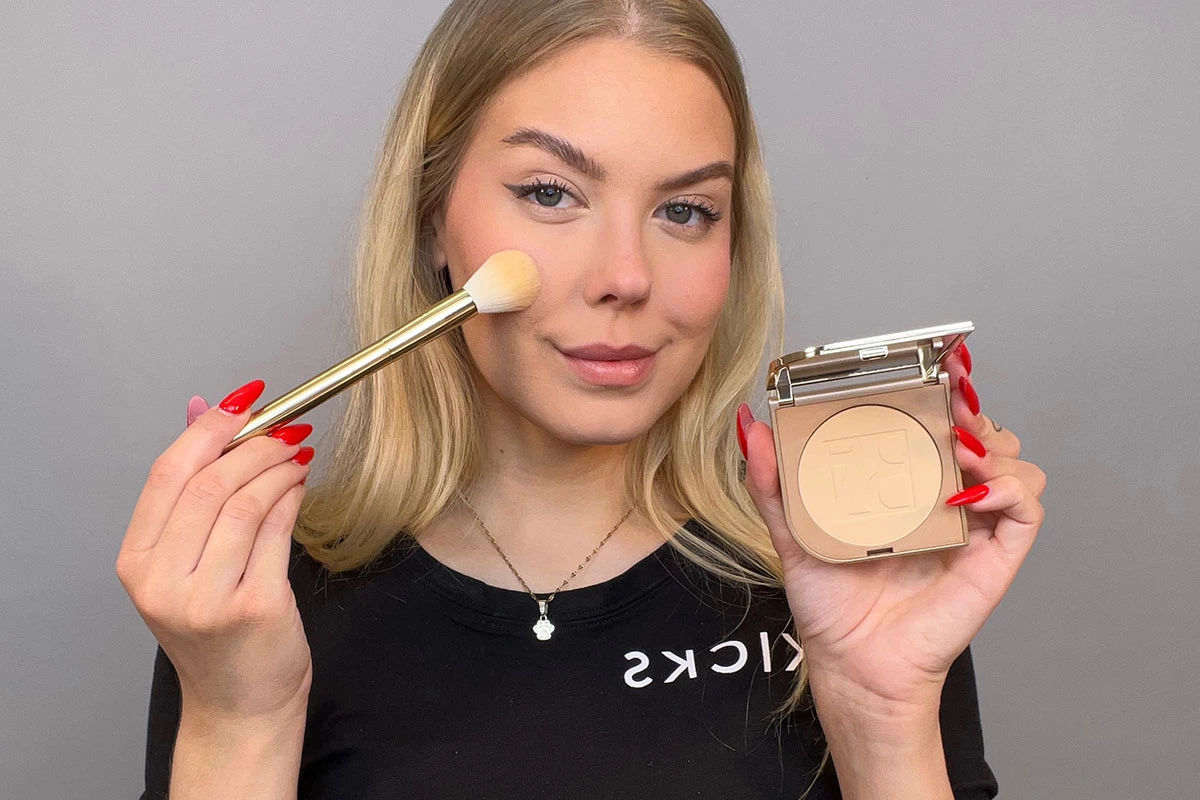 En närbild på en person med långt blont hår som håller i en sminkborste och en öppen puderkompakt mot en grå bakgrund.