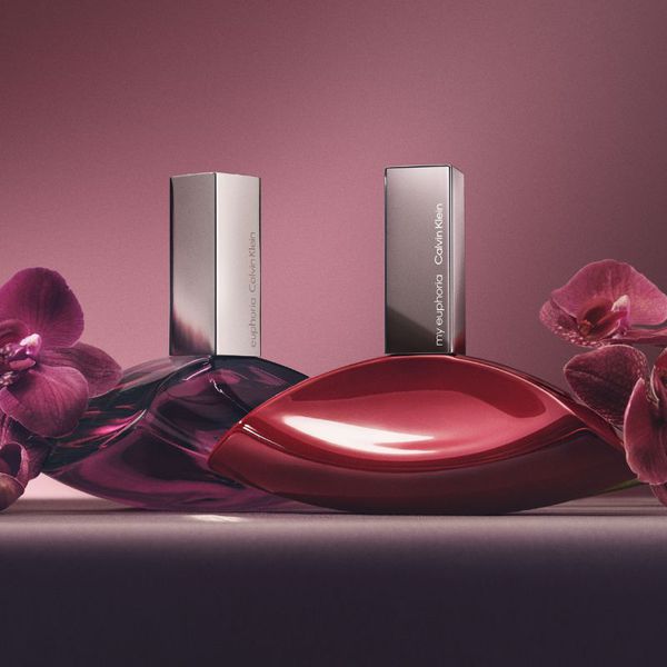 Två parfymer från Calvin Klein med namnen "euphoria" och "my euphoria". Flaskorna har en elegant, böjd design i röd och lila färg, med silverfärgade korkar. De är placerade på en yta med en rosa bakgrund och omgivna av lila orkidéer.