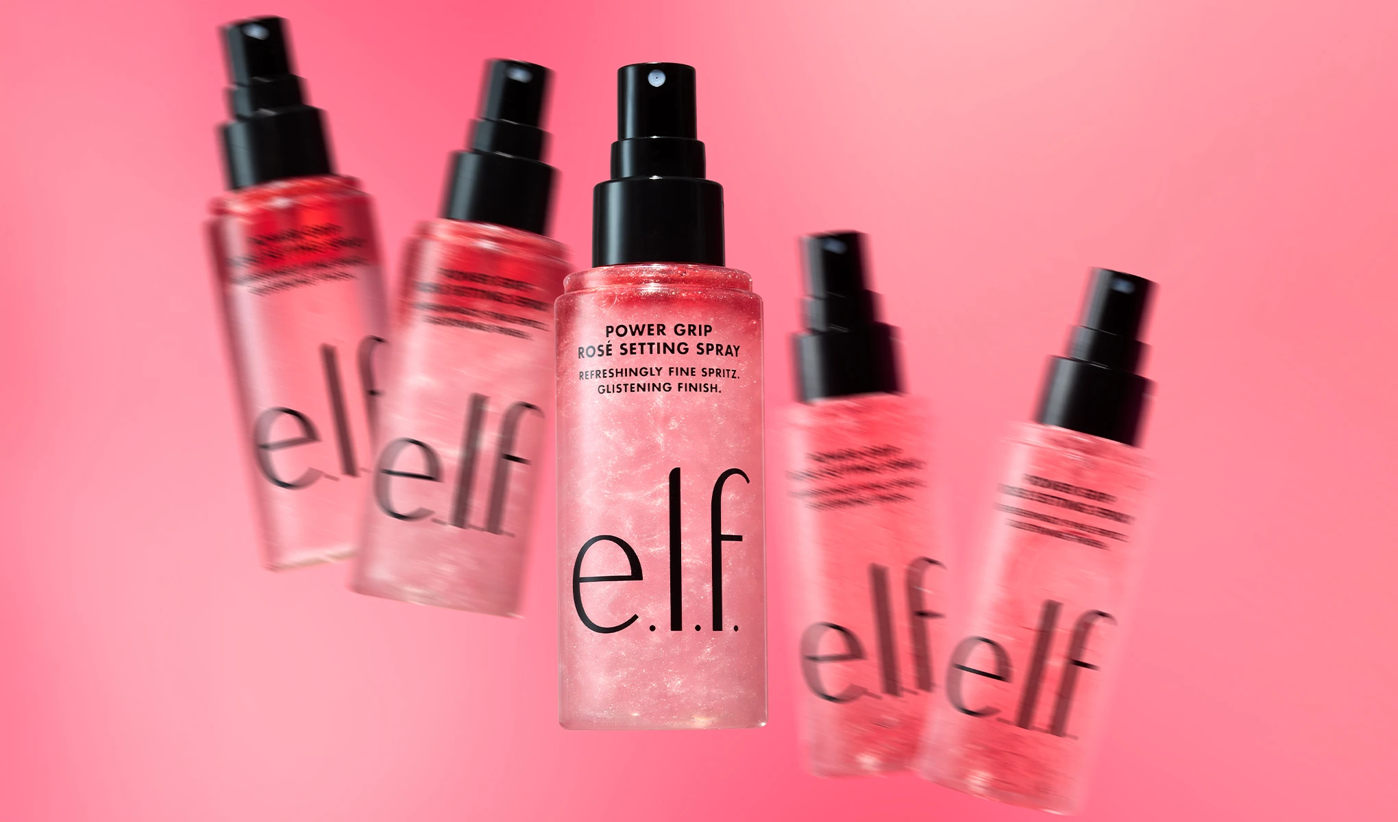 Useita e.l.f. Power Grip Rosé Setting Spray -meikinkiinnityssuihkeita näkyy vaaleanpunaista taustaa vasten, ja yksi pullo on keskellä tarkennettuna. Pullossa näkyvät tekstit "POWER GRIP ROSÉ SETTING SPRAY", "REFRESHINGLY FINE SPRITZ.", "GLISTENING FINISH." sekä tuotemerkki "e.l.f.", mikä korostaa hohtavaa viimeistelyä antavaa meikinkiinnityssuihketta.