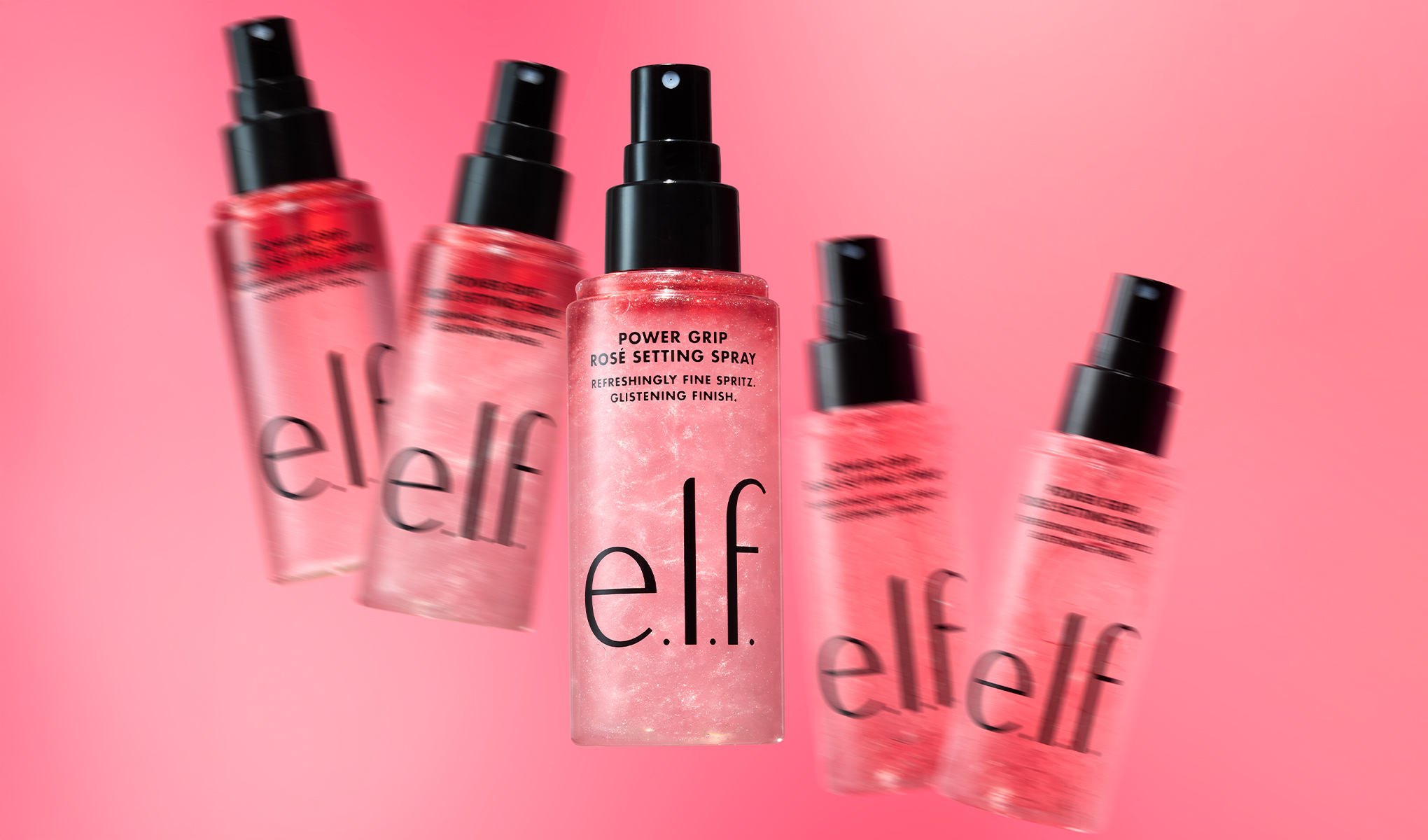 Flera flaskor av e.l.f. Power Grip Rosé Setting Spray visas mot en rosa bakgrund, med en produkt i fokus i mitten. På flaskan syns texten "POWER GRIP ROSÉ SETTING SPRAY", "REFRESHINGLY FINE SPRITZ." , "GLISTENING FINISH." och varumärket "e.l.f.", vilket framhäver en fixerande makeupspray med skimrande finish.