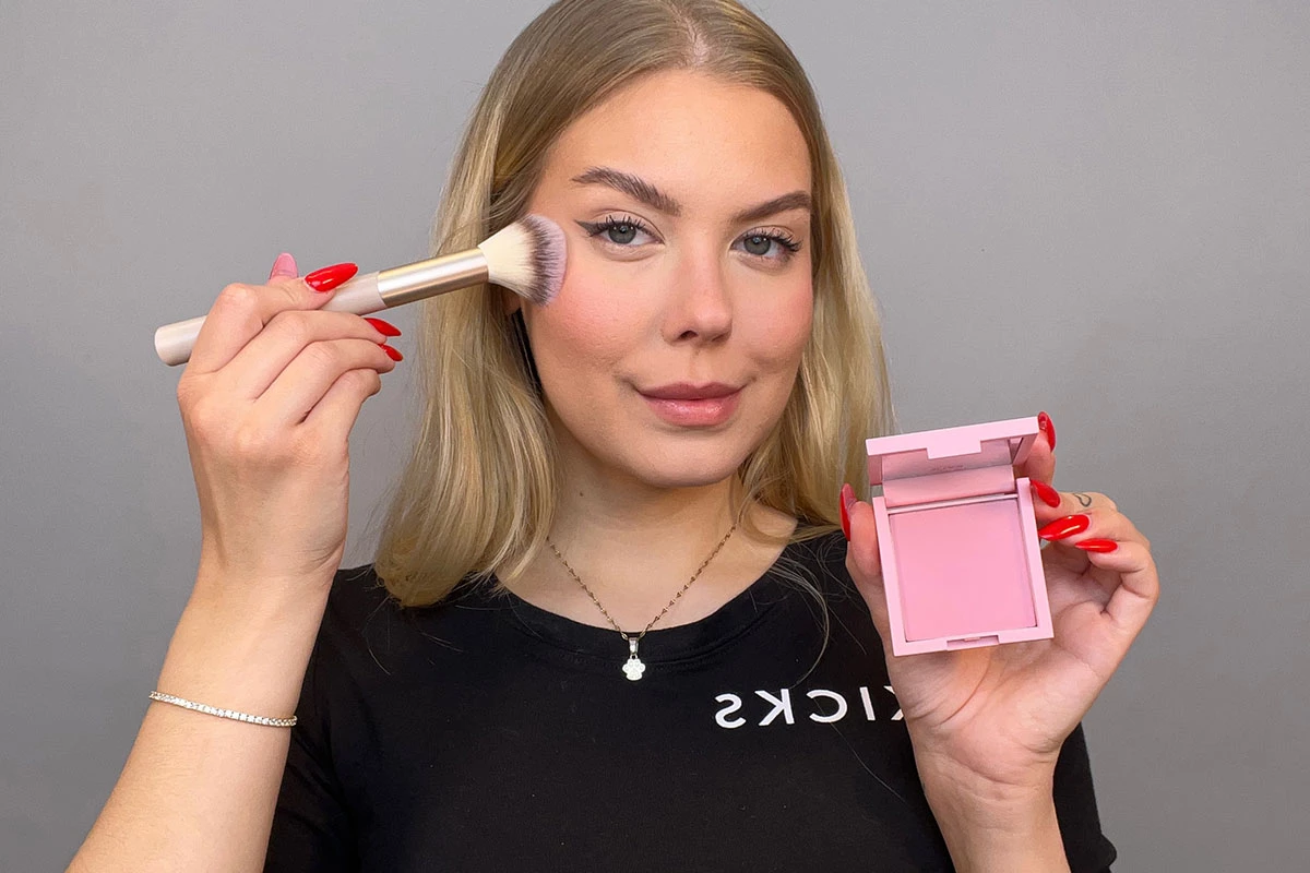 En närbild på en person med långt blont hår som håller i en sminkborste och en öppen rosa sminkpalett mot en grå bakgrund.