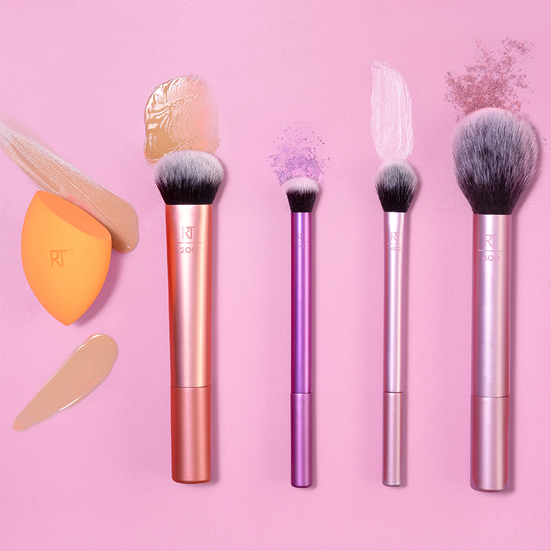 Fyra sminkborstar och en orange sminksvamp från Real Techniques är arrangerade mot en rosa bakgrund. Borstarna har skaft i färgerna roséguld, lila och silver, och svarta borst. Runt borstarna finns olika sminkprodukter applicerade, inklusive foundation, puder och kräm. Sminksvampen har foundation på sig och ligger till vänster i bilden.
