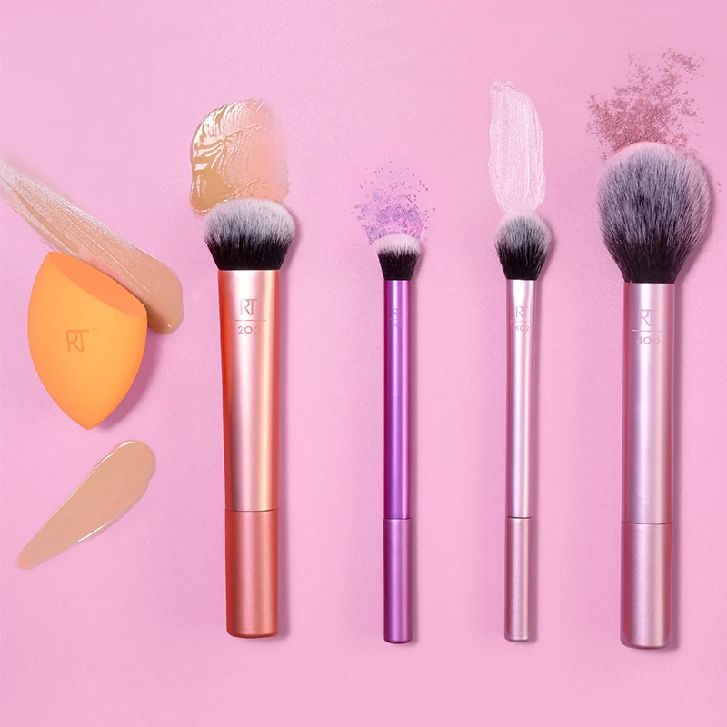 Fyra sminkborstar och en orange sminksvamp från Real Techniques är arrangerade mot en rosa bakgrund. Borstarna har skaft i färgerna roséguld, lila och silver, och svarta borst. Runt borstarna finns olika sminkprodukter applicerade, inklusive foundation, puder och kräm. Sminksvampen har foundation på sig och ligger till vänster i bilden.