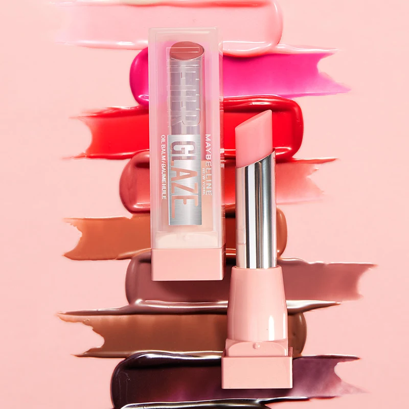 Maybelline-huulituote ja pakkaus vaaleanpunaisella taustalla, ympärillä levitettyjä huulivärisävyjä.