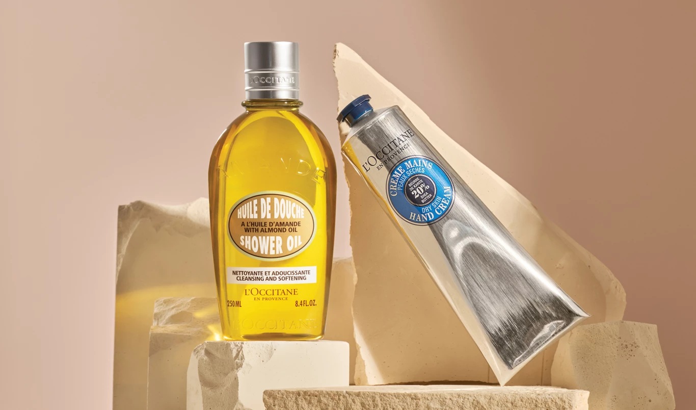 To produkter fra L'Occitane. Den ene er en flaske og den andre er en tube. Produktene er plassert på beige steiner med beige bakgrunn.