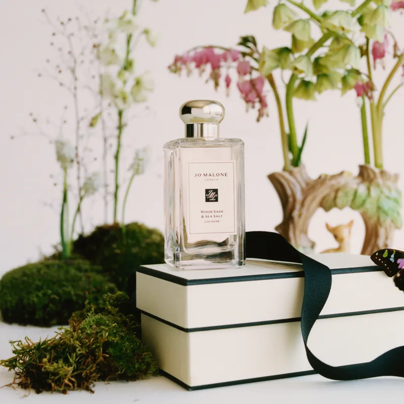 En parfymflaska från Jo Malone med texten "Wood Sage & Sea Salt Cologne" placerad på en vit presentask med svart band. Bakgrunden är dekorerad med gröna växter, mossa och blommor.