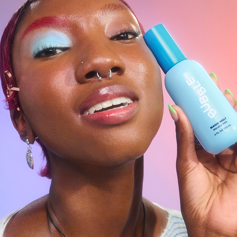 Henkilö pitää kädessään Bubble Magic Melt meikinpoistosuihketta vaaleansinisessä pullossa. Tausta on värikäs, vaaleanpunaisen ja violetin sävyissä.