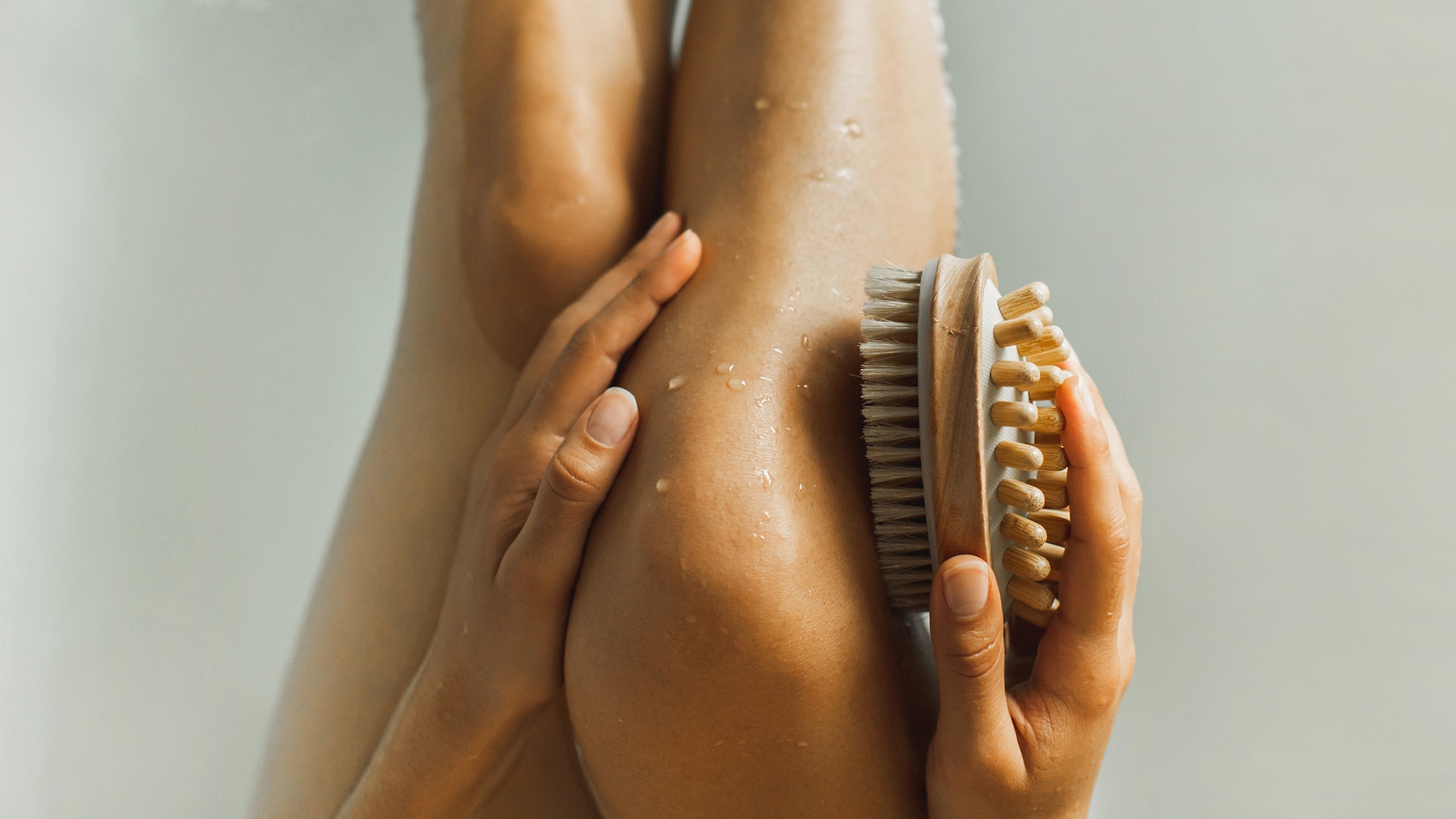 Henkilö käyttää kuivaharjaa vartalonhoitoon jalalla, jonka iholla näkyy vesipisaroita. Puinen harja luonnonvärisillä harjaksilla pidetään jalkaa vasten minimalistisessa spa- ja wellness-ympäristössä, jossa korostuvat kuorinta ja body care.
