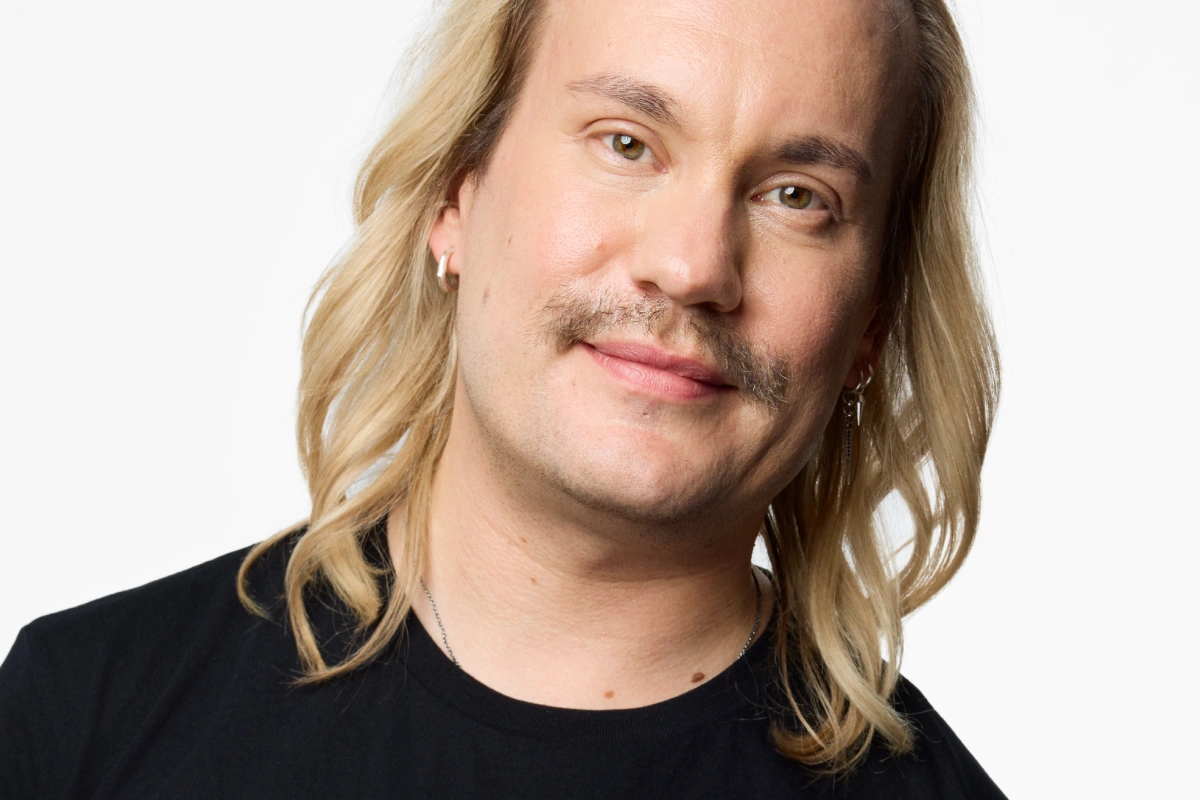 Portrett av en person med langt lyst hår og bart, iført svart genser, mot lys bakgrunn.