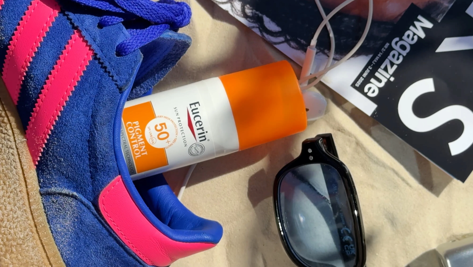 Blå sneaker, Eucerin Sun Protection Pigment Control SPF 50+, solbriller og magasin på sand.