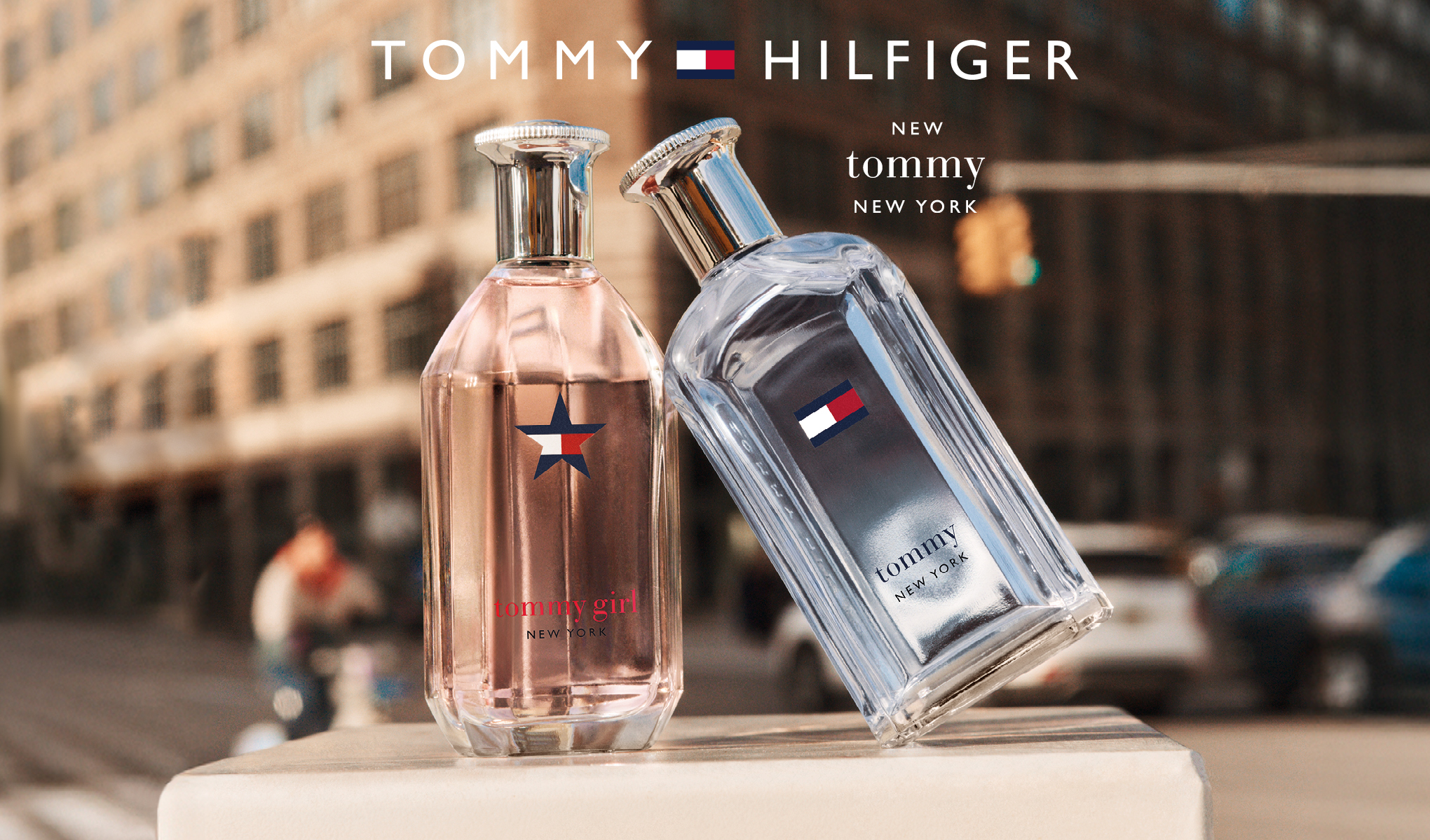 Tommy Hilfiger dofter. Två flaskor, en rosa och en klar, står utomhus i en stadsmiljö. På den rosa flaskan står det "tommy girl NEW YORK" och på den klara flaskan står det "tommy NEW YORK". Båda flaskorna har Tommy Hilfiger-logotypen.
