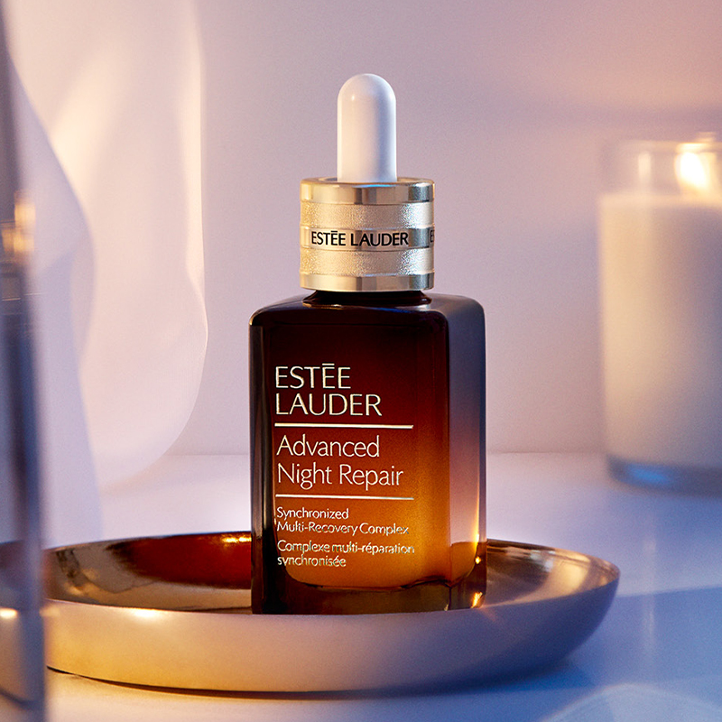 Pullo Estée Lauderin Advanced Night Repair Synchronised Multi-Recovery Complex -yövoiteen valmistetta, joka on asetettu kultaiselle lautaselle valoisaa taustaa vasten, ja taustalla on valaistu lyhty.