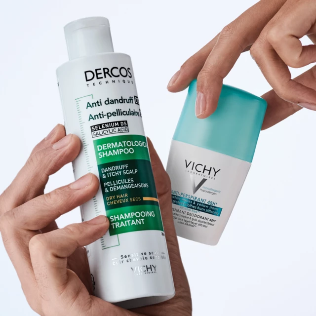 Två händer som håller upp en shampooflaska från Dercos och en anti-persiprant produkt från Vichy mot en ljusblå vägg.